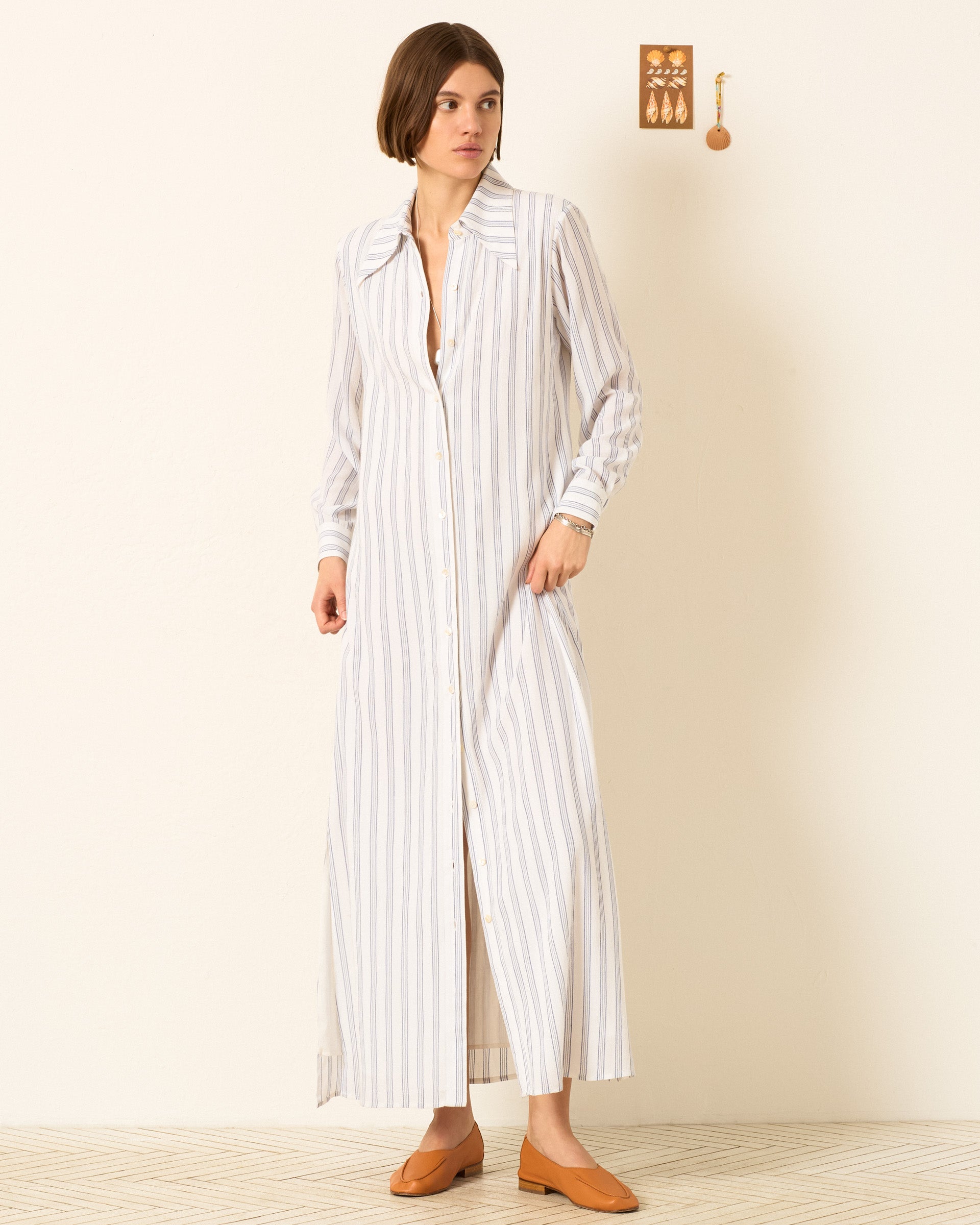 A woman stands indoors wearing the Lotte Coastal Stripe Dress, a long, striped shirt dress featuring a '70s dagger collar and mother-of-pearl buttons, paired with brown loafers. A small wall decoration is visible in the background.