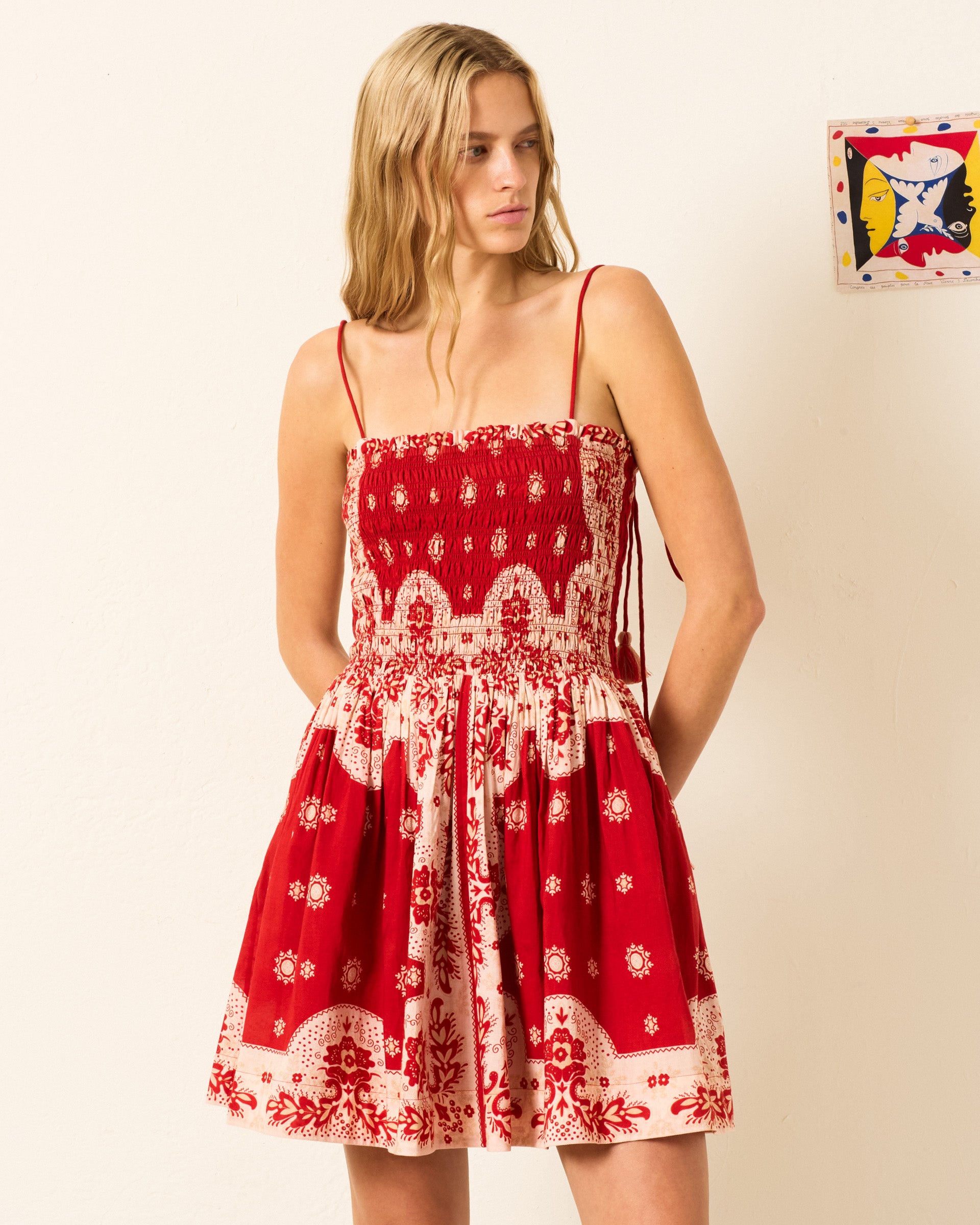 A woman wearing the Merida Lava Bandana Dress, a red and white bandana-print summer dress with thin straps, stands against a light-colored wall looking to the side.