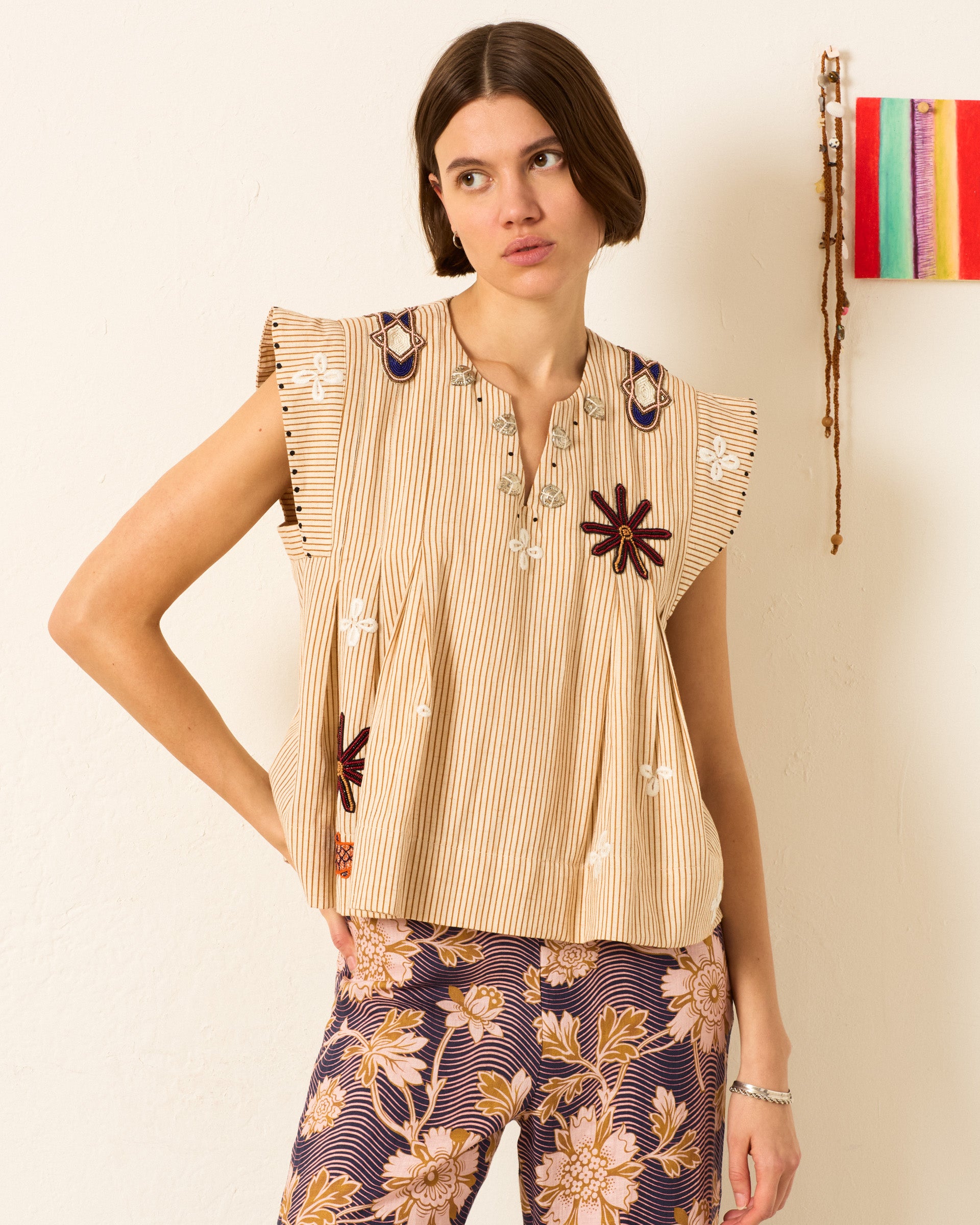 A woman stands against a white wall wearing the Nadine Cedar Coin Top, a hand-embroidered beige striped blouse, paired with wide-leg floral pants in purple and beige tones.