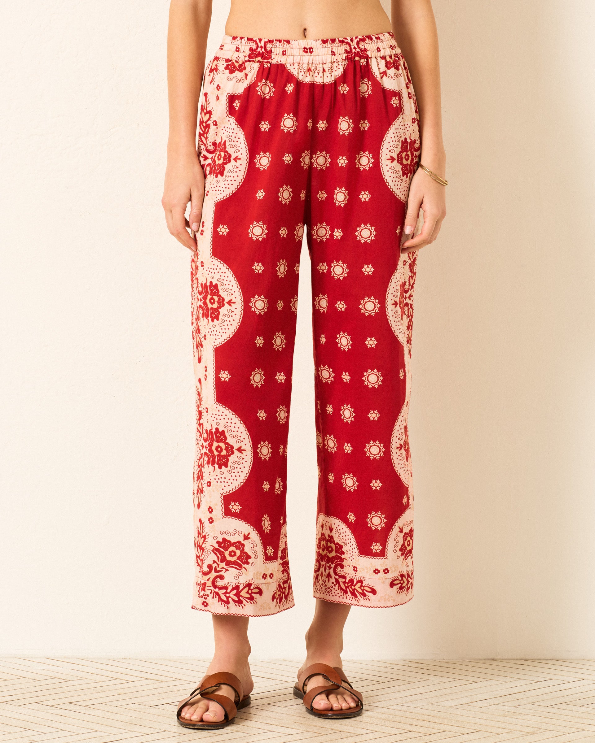 Someone stands against a light background wearing the Otto Lava Bandana Pant—red and cream, bandana-print, wide-leg pants with an elastic waistband—paired with brown sandals.