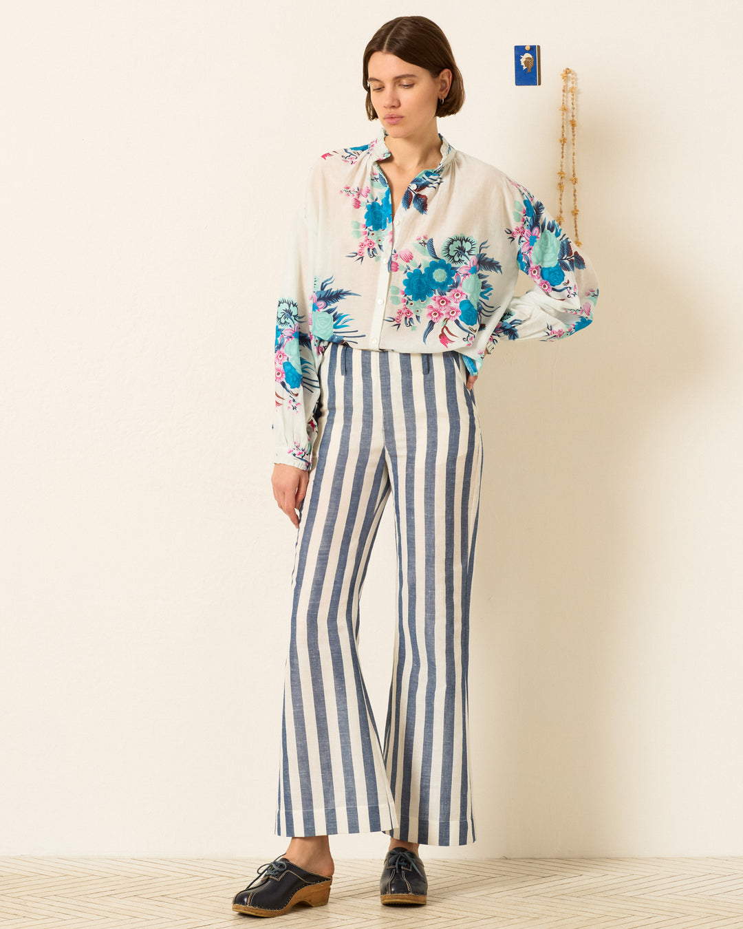 A woman stands indoors wearing the Poet Juneberry Blouse, a vintage-inspired top with a ruffled collar, paired with high-waisted blue-and-white striped pants and dark oxford shoes.