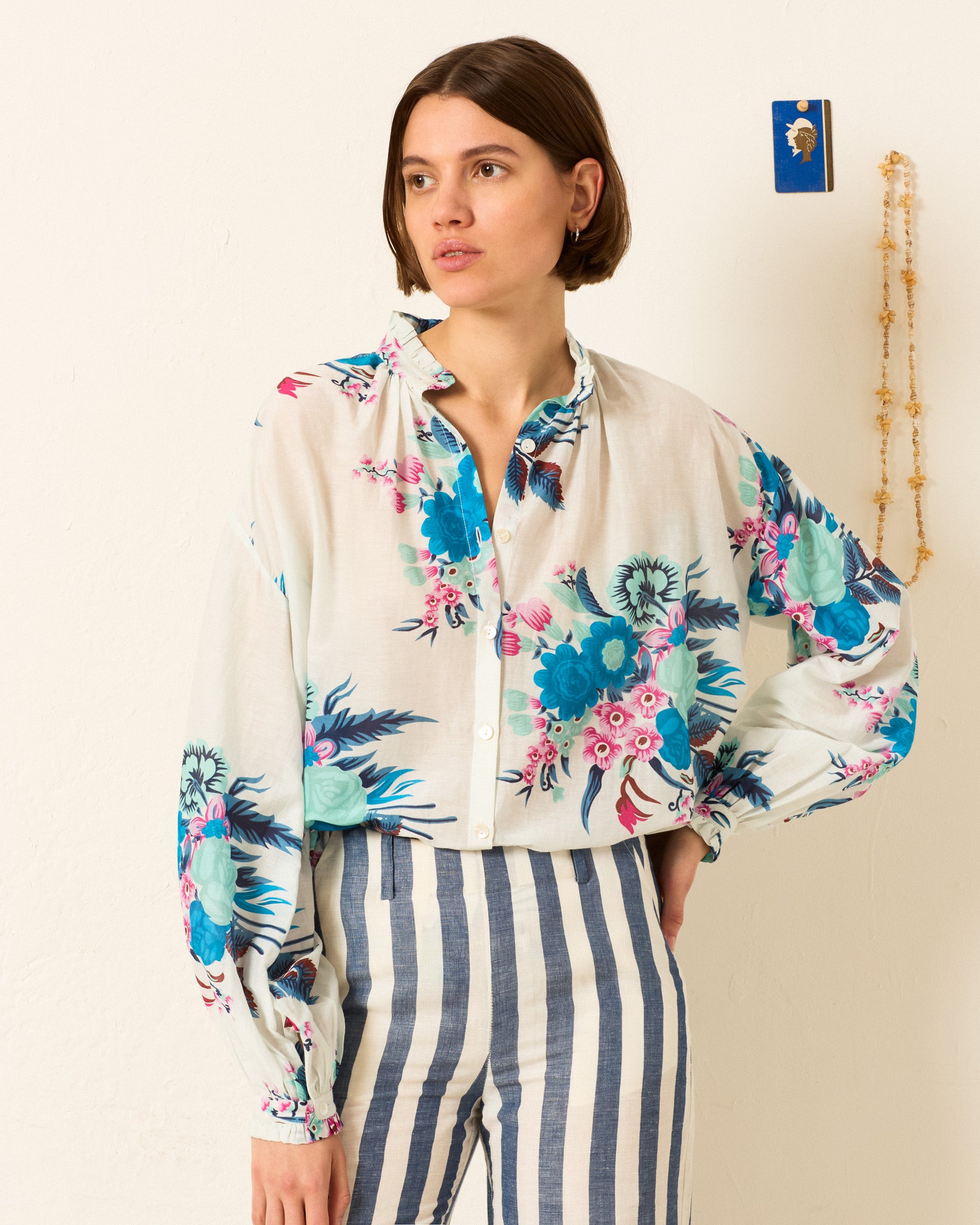 A woman stands against a plain wall wearing the Poet Juneberry Blouse, a vintage-inspired cotton-silk shirt with blue and pink floral patterns, paired with high-waisted, blue-and-white striped pants.