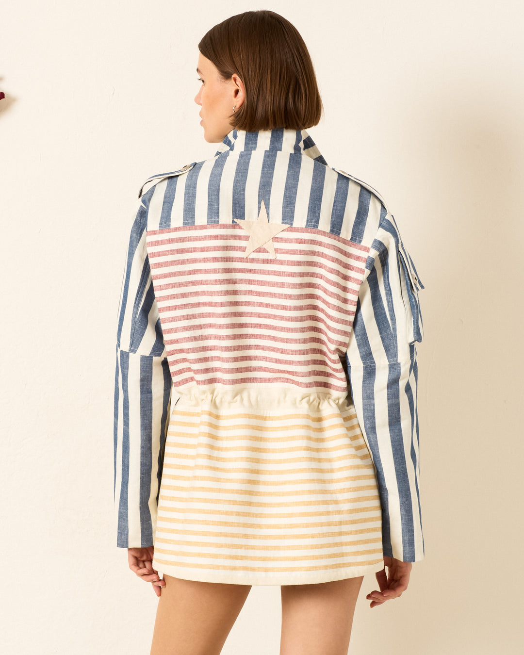 A woman faces away, wearing the Porter Sailor Stripe Jacket featuring blue and white vertical stripes, red and white horizontal stripes, a beige striped lower section, and a handmade star appliqué on the back.