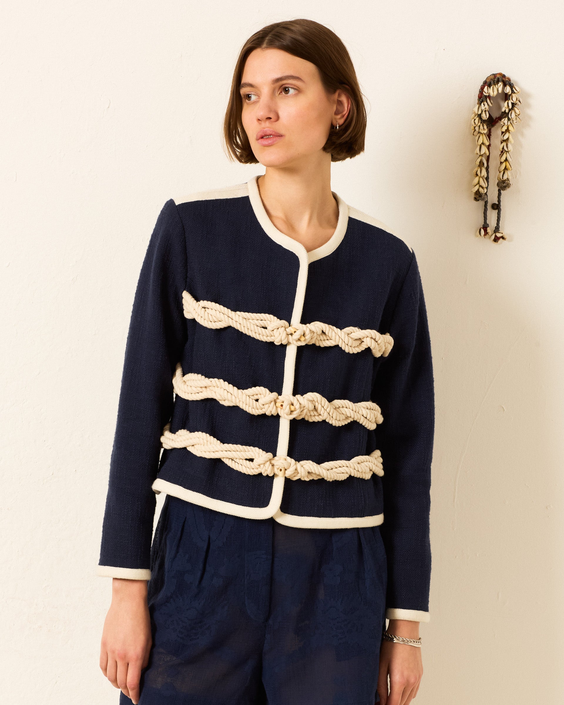 A woman stands by a plain wall in the Rowan Deep Sea Jacket, featuring cream rope detailing, paired with dark patterned pants. A shell-adorned decorative object hangs beside her.
