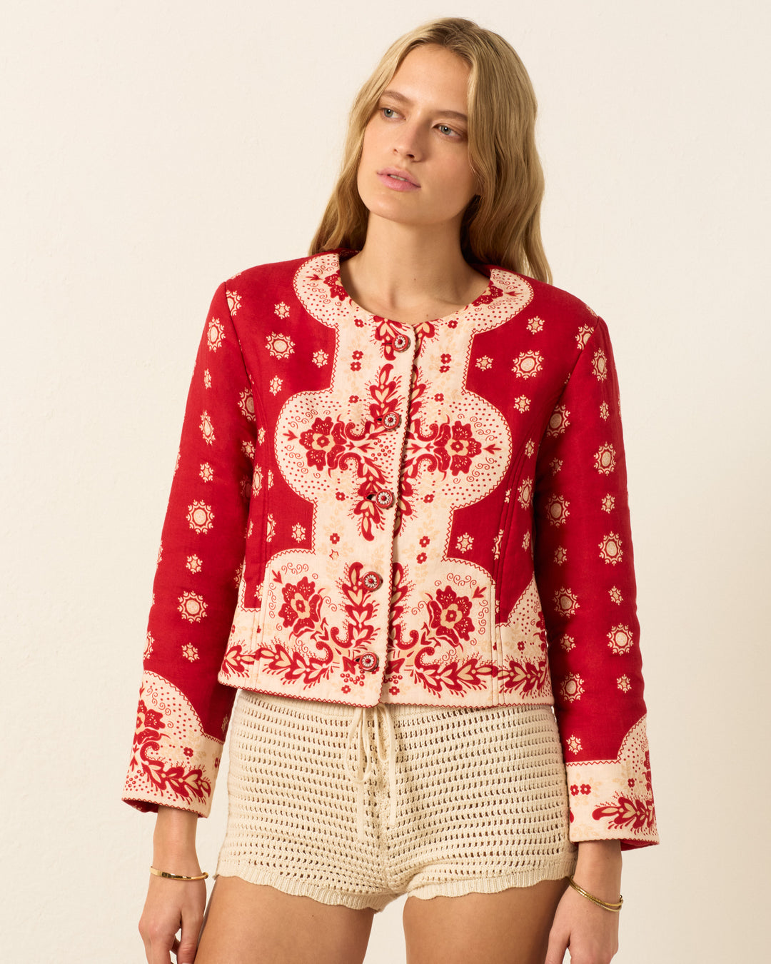 A woman stands against a plain light background wearing the Rowan Lava Bandana Jacket, featuring a red and cream bandana print, long sleeves, and handmade buttons, paired with cream crochet shorts.