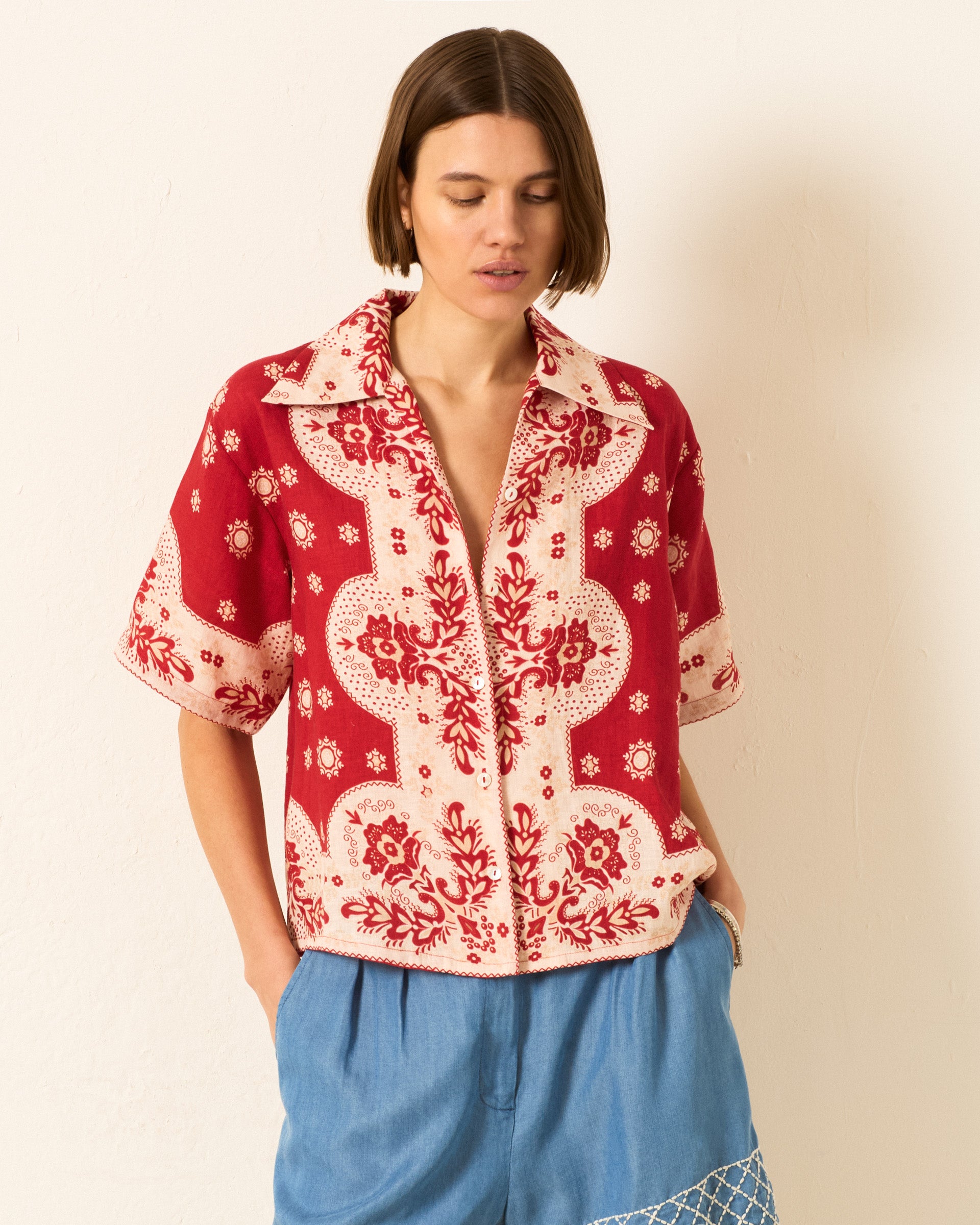 A woman wears the Stevie Lava Bandana Shirt, a vintage Hawaiian top with a red and white bandana print, paired with loose blue pants, as she stands against a plain light-colored wall.