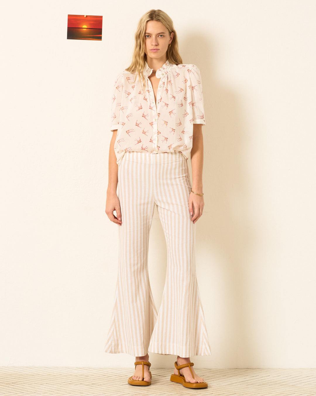 A woman stands indoors wearing the Winnie Cedar Swallow Shirt, a hand-block-printed cotton-silk blend with a ruffled collar, paired with high-waisted striped flared pants and sandals. A small sunset photo hangs on the wall behind her.