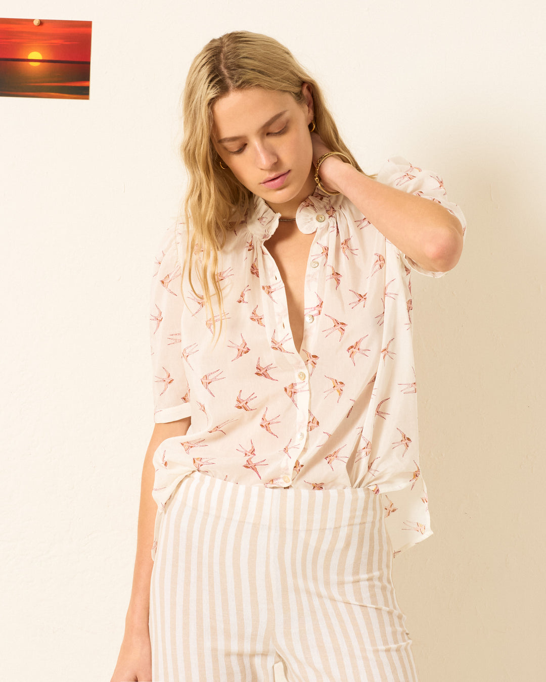 A person with long blond hair wears the Winnie Cedar Swallow Shirt, a hand-block-printed white button-up with a bird pattern, paired with white striped pants and standing before a neutral wall featuring a small sunset photo.
