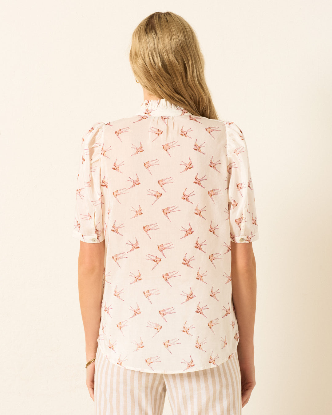 A person with long blonde hair is seen from behind, wearing the Winnie Cedar Swallow Shirt, a hand-block-printed cotton-silk blend top featuring a bird print, paired with light striped pants against a plain background.