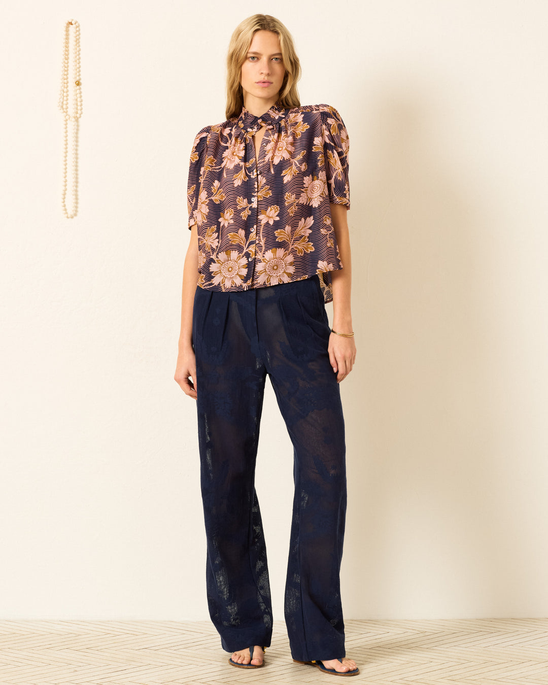 A person stands indoors in the Winn Wavy Zinnia Shirt, featuring a floral motif and short sleeves, paired with navy blue sheer pants and black sandals against a plain wall.