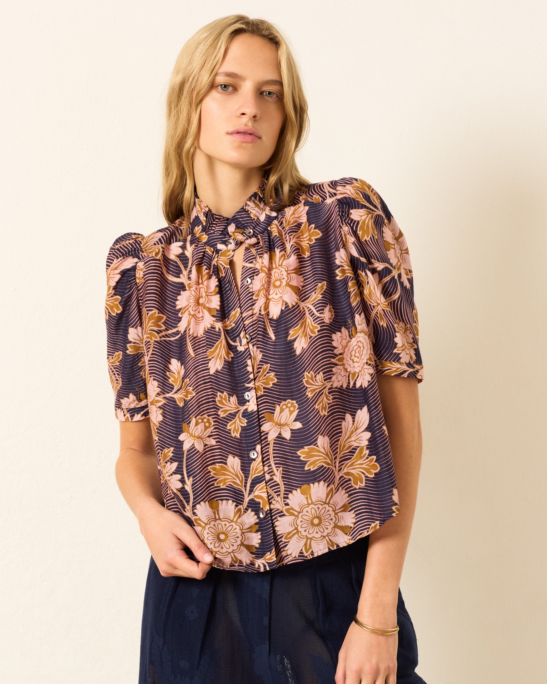 A woman wears the Winn Wavy Zinnia Shirt, a short-sleeve, button-up blouse in a cotton-silk blend with a purple and pink floral print, standing against a plain light wall.