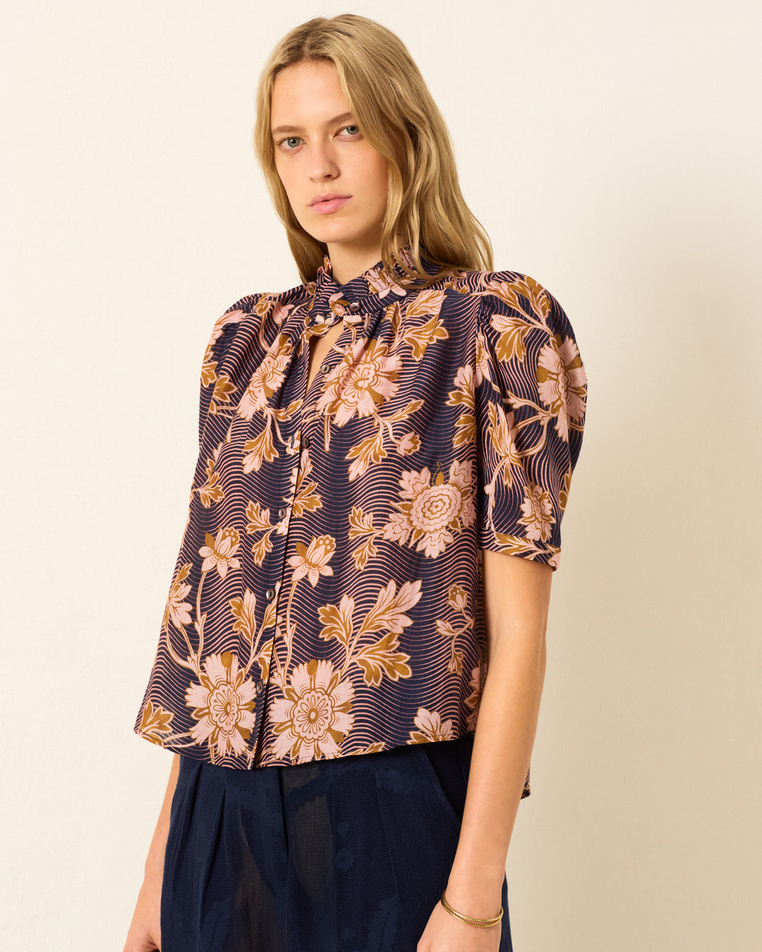 A woman wears the Winn Wavy Zinnia Shirt, a short-sleeved button-up blouse with puff shoulders and a purple and orange floral motif, paired with navy pants against a plain light background.