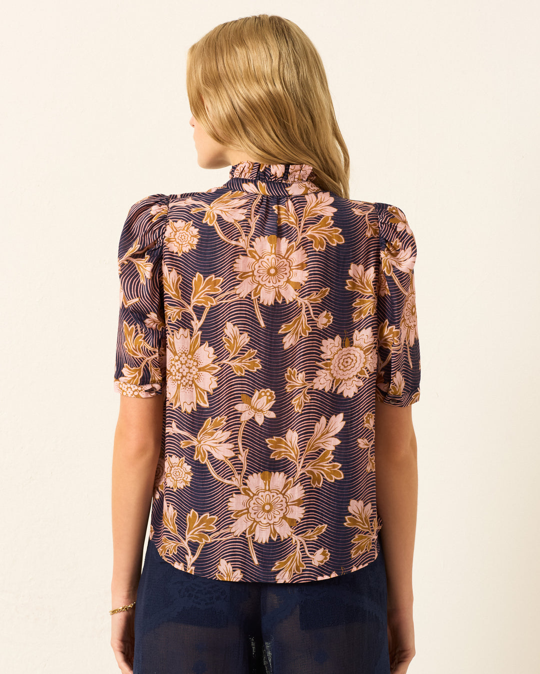 A woman with blonde hair is seen from the back wearing the Winn Wavy Zinnia Shirt, a navy cotton-silk blouse featuring a pink and orange floral motif and short puff sleeves.