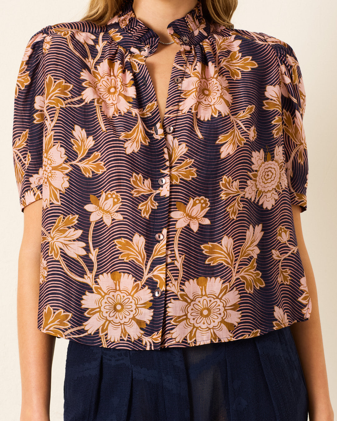 A woman wearing the Winn Wavy Zinnia Shirt, a short-sleeved, button-up blouse with puff shoulders and a navy base featuring a pink and beige floral print, paired with a dark skirt.