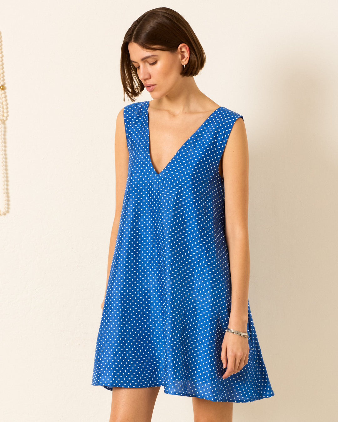 A woman wears the Soleil Harbor Dot Silk Dress, a sleeveless blue dress with white polka dots, standing against a plain light-colored wall.
