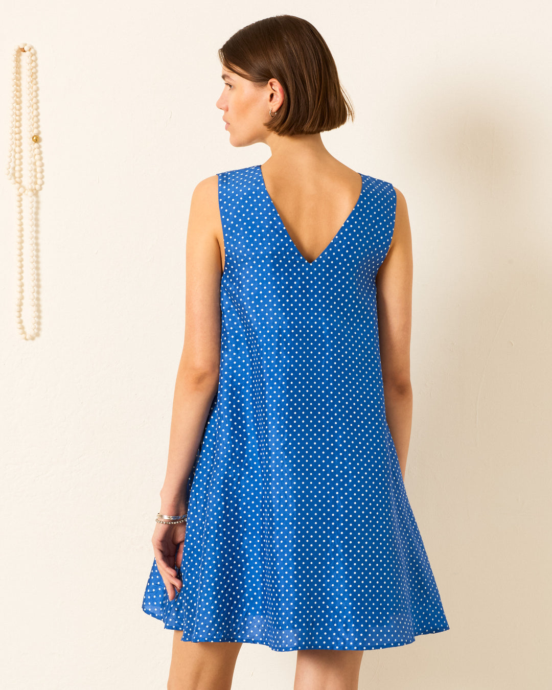 A woman stands facing away in the Soleil Harbor Dot Silk Dress, a sleeveless blue trapeze style with white polka dots and a V-shaped back; a string of pearls hangs on the wall to her left.