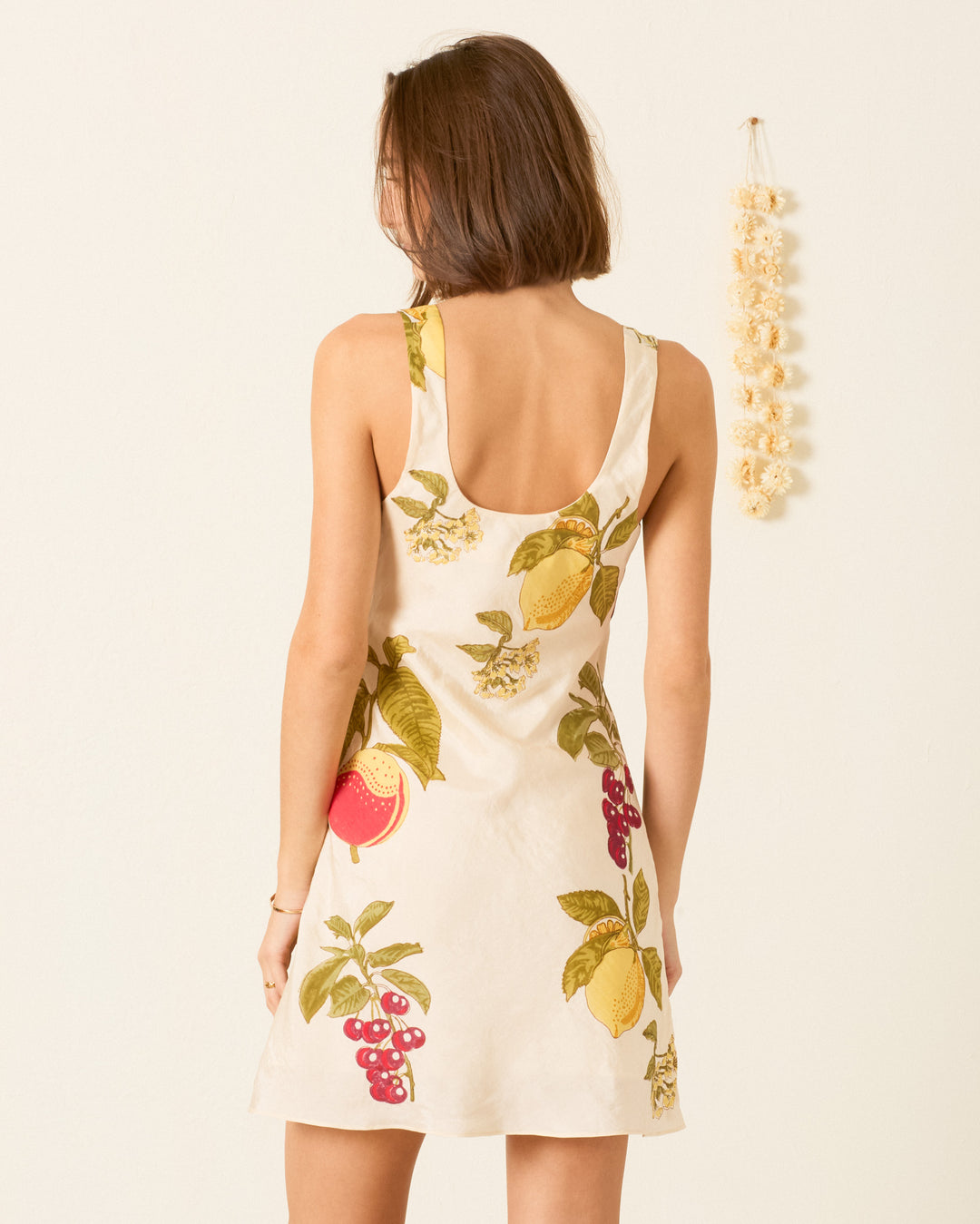 A woman with short brown hair stands with her back to the camera, wearing the Addie Four Fruit Silk Dress—an artisan mini dress featuring a fruit print—near a beige wall decorated with a hanging ornament.