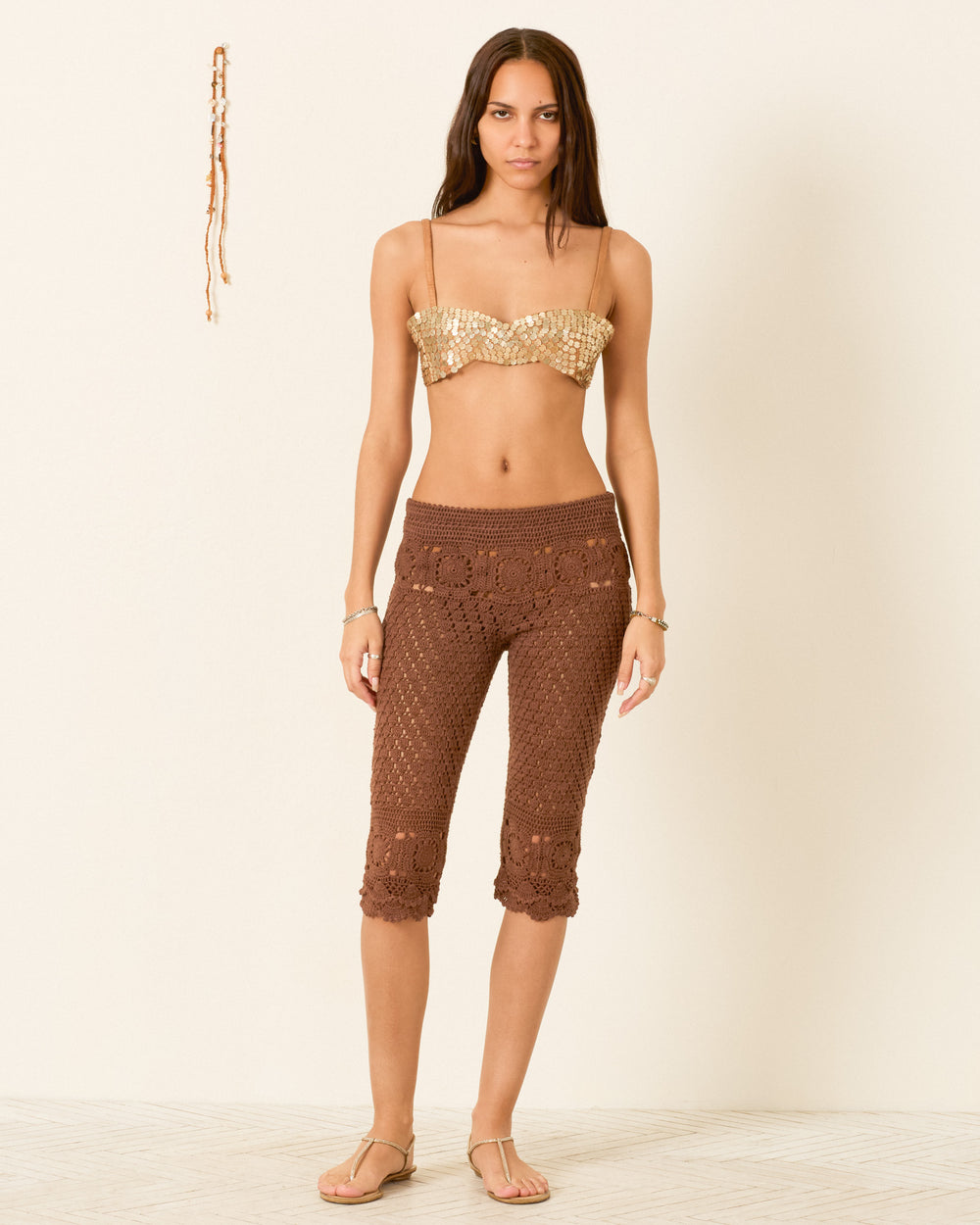 A woman stands against a plain wall wearing a gold sequin bandeau, Aletta Clove Mandala Pant in brown openwork crochet, and sandals. A beaded accessory hangs on the wall beside her.