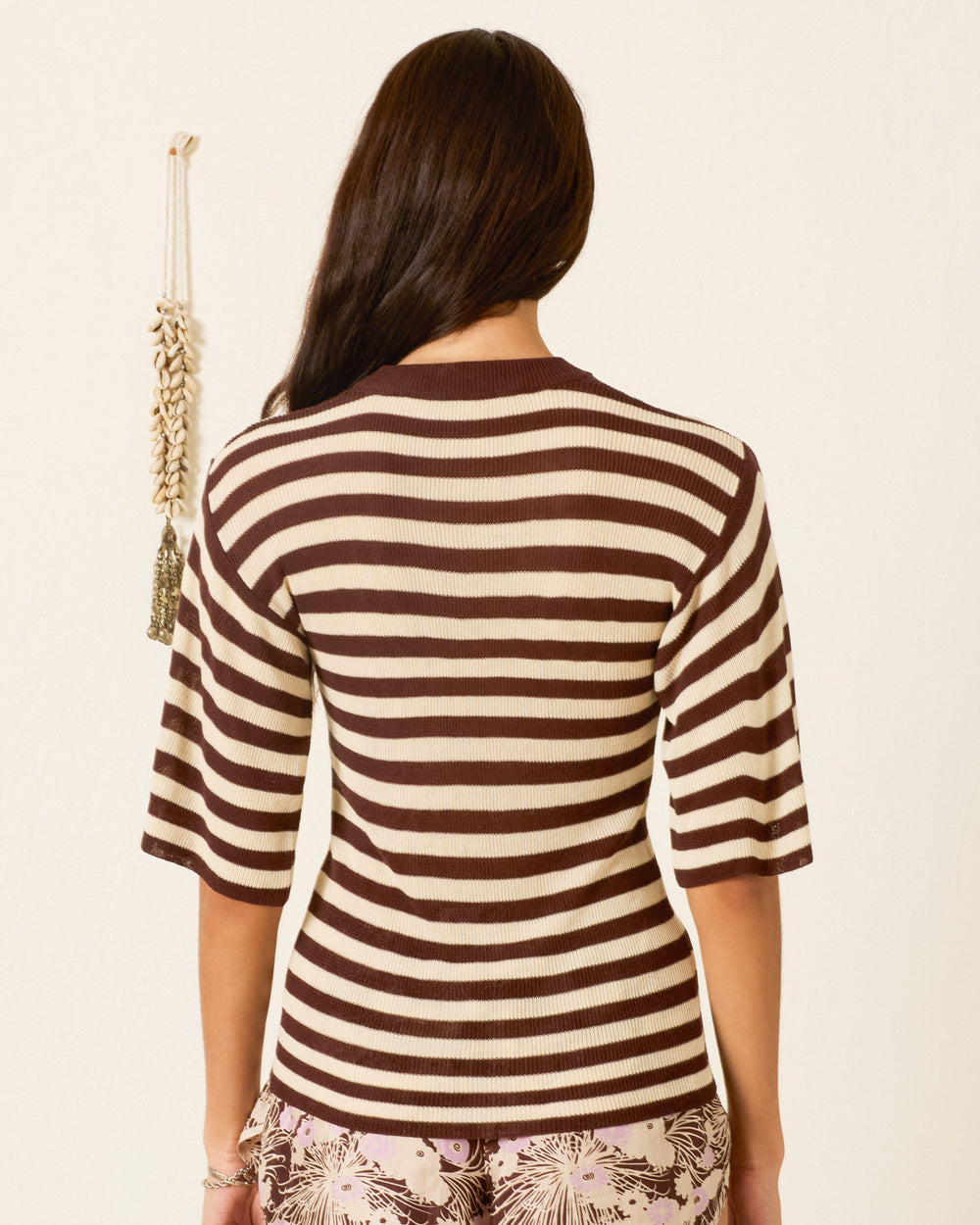 Someone with long dark hair stands with their back to the camera, wearing the Aless Clove Stripe Top—a brown and white striped sweater crafted from soft bamboo viscose.