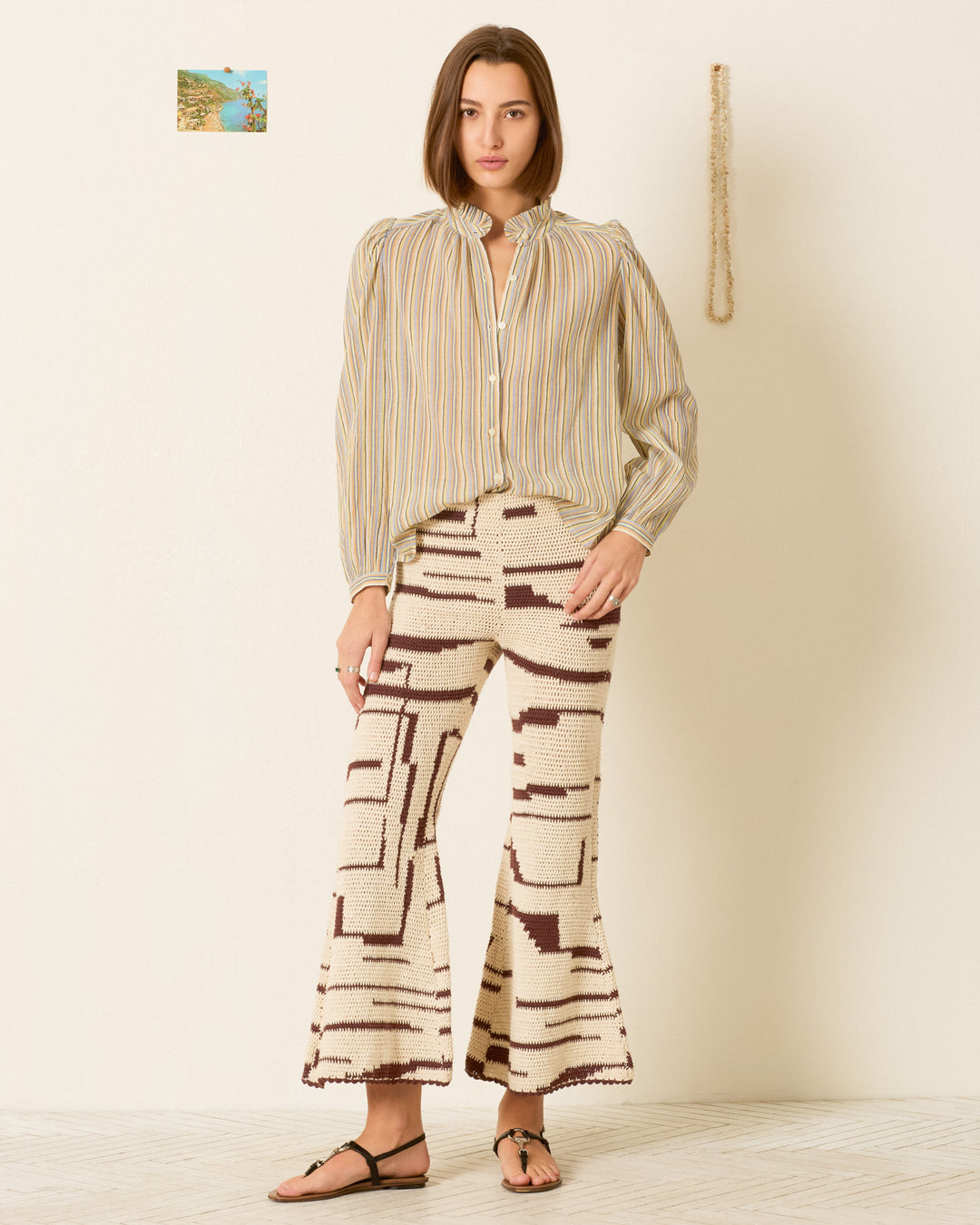 A woman stands indoors wearing the Annabel Sandlilt Stripe Shirt, patterned wide-leg pants, and sandals. A small picture and rope hang on the wall behind her.