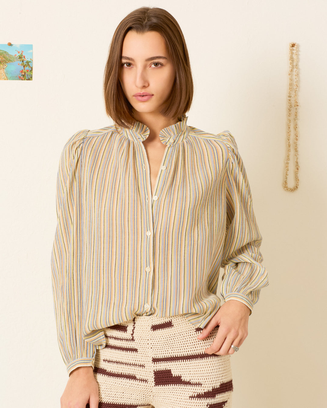 A woman with straight brown hair wears the Annabel Sandlilt Stripe Shirt, standing against a neutral wall with a small photo and hanging decoration in the background.
