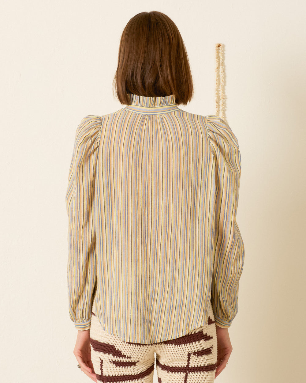 Wearing the Annabel Sandlilt Stripe Shirt, a person with shoulder-length brown hair stands with their back to the camera against a plain light wall. The long-sleeve, high-neck blouse features stripes and subtle puff shoulders.
