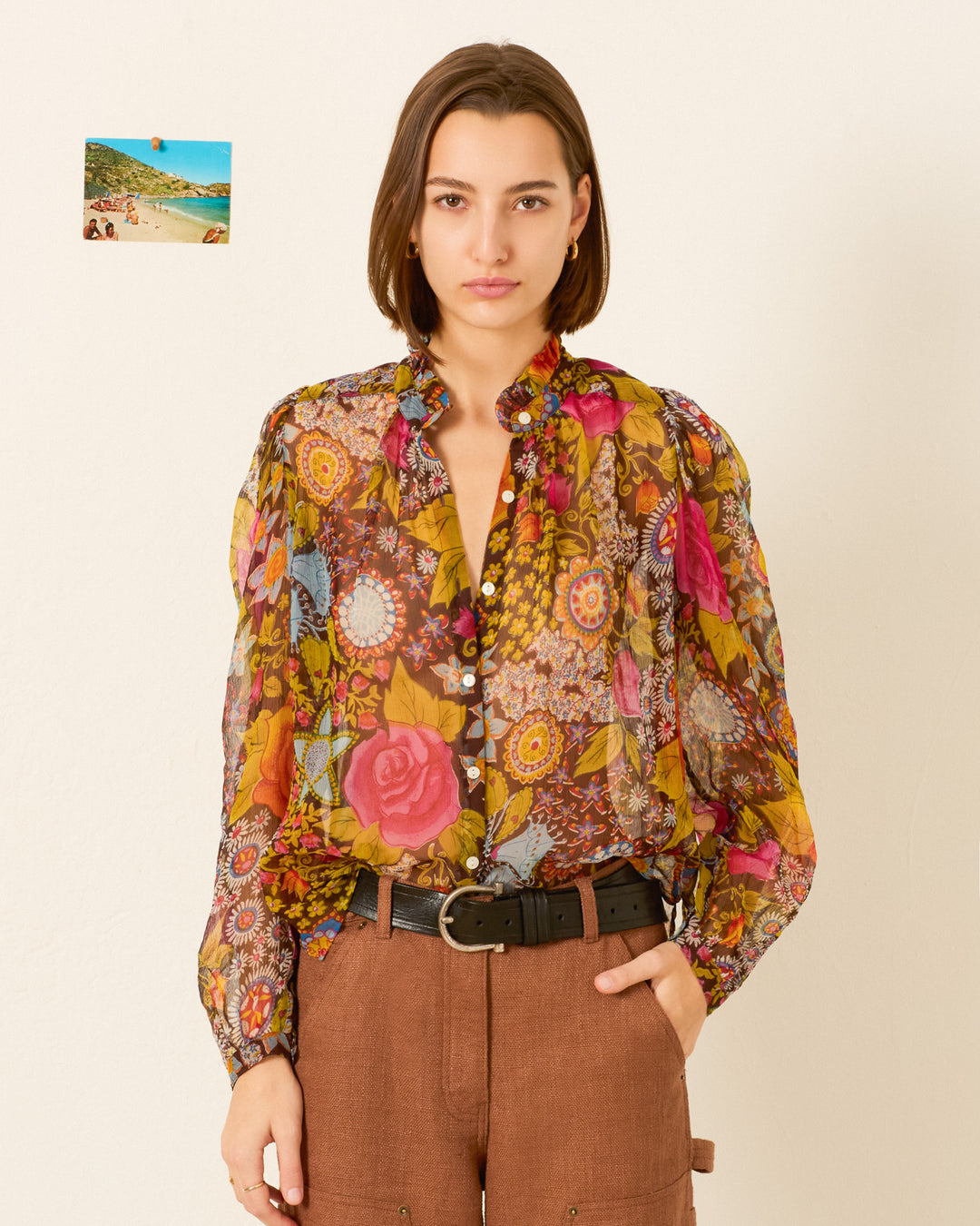 A woman stands against a white wall wearing the Annabel Bloom Mirage Shirt, a psychedelic floral blouse with a ruffled collar, paired with brown pants and a black belt; a small photo is pinned to the wall behind her.
