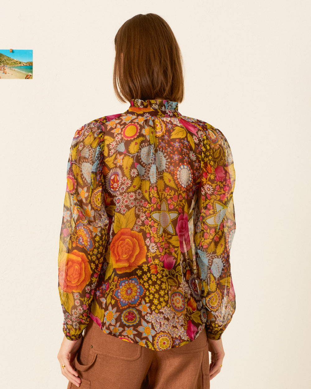 Wearing the Annabel Bloom Mirage Shirt—a sheer, long-sleeved blouse with puff shoulders and psychedelic floral prints—a person with straight brown hair and brown pants stands with their back to the camera.