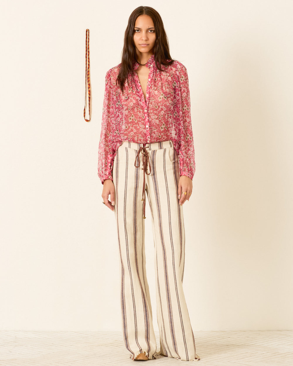 Woman standing indoors, wearing the Annabel Berry Cosmos Shirt with high-waisted, wide-leg striped pants; a patterned scarf hangs on the wall behind her.