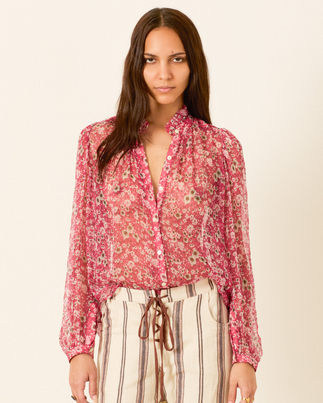 A woman wears the Annabel Berry Cosmos Shirt, a vintage floral top with sheer pink fabric, buttons, and long sleeves, paired with high-waisted beige pants with vertical brown stripes.