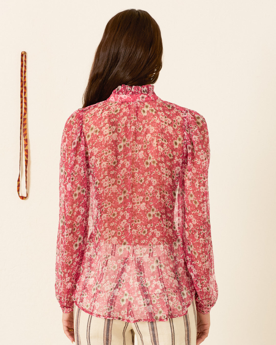 A woman with long brown hair, seen from behind, wears the Annabel Berry Cosmos Shirt—sheer with a vintage floral pattern and long sleeves—paired with striped pants. A thin patterned belt hangs on the wall to her left.