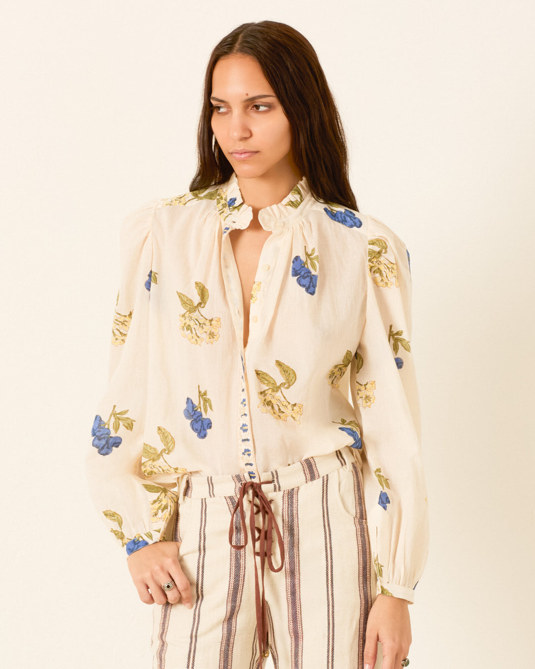 A woman stands against a plain background, looking to the side, wearing the Annabel Blueberry Flora Shirt—a hand block printed cream blouse with blue and yellow floral prints—paired with striped pants.