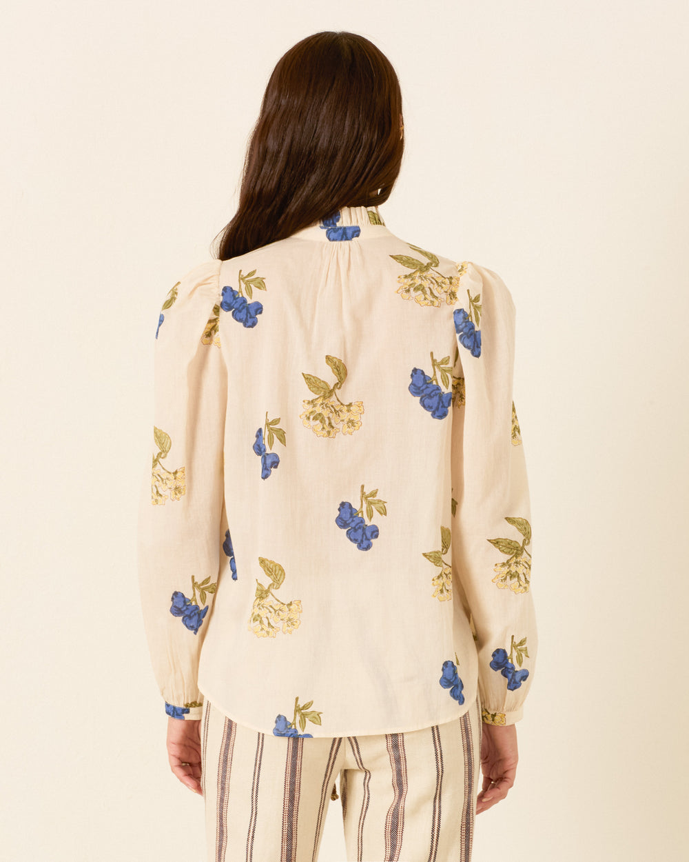A person with long brown hair is shown from the back wearing the Annabel Blueberry Flora Shirt, a hand block printed cream blouse with blue and yellow floral prints, paired with striped pants against a plain light background.