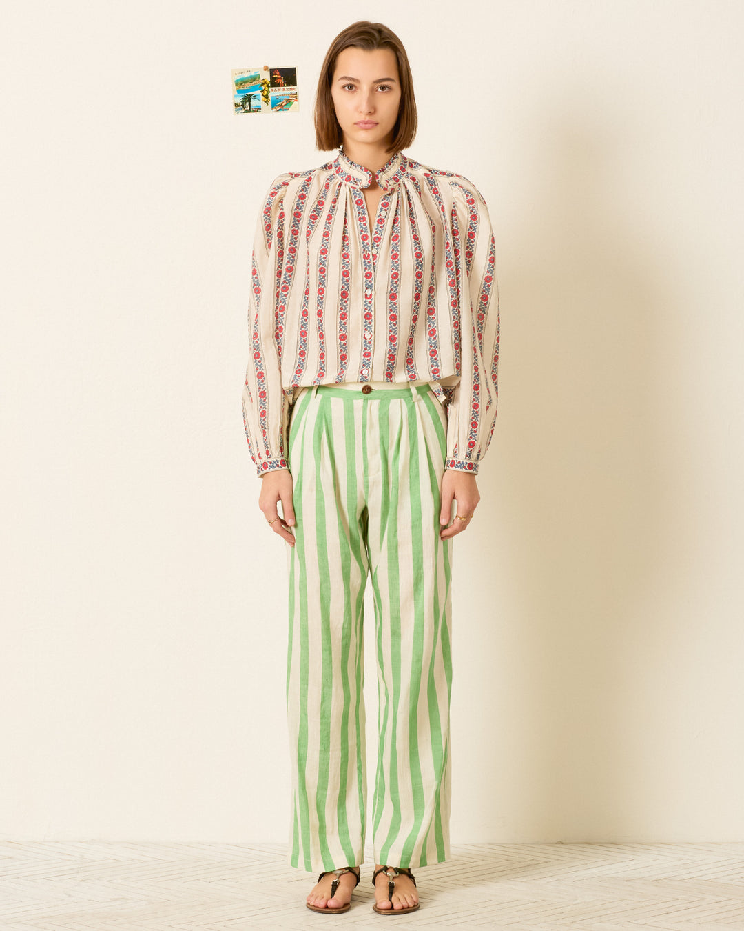 A person stands against a plain wall wearing the Annabel Roseband Stripe Shirt, styled with green and white striped pants and sandals. Two small photos are on the wall behind.