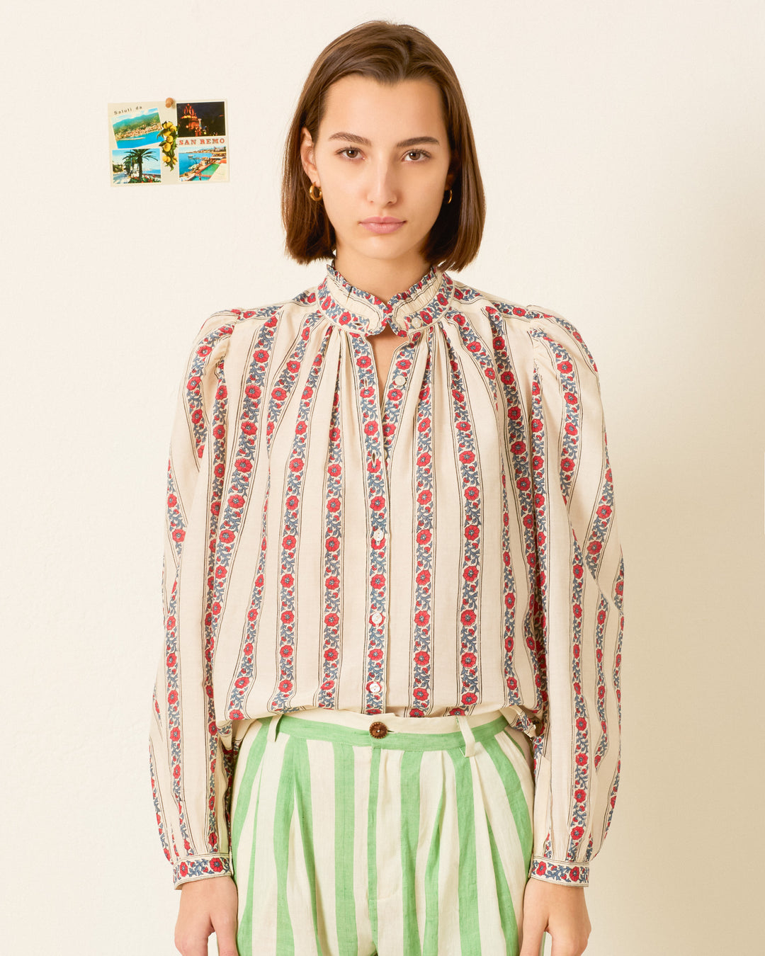 A person stands indoors wearing the Annabel Roseband Stripe Shirt, a hand-block-printed floral striped blouse with a ruffled collar, paired with green striped pants. Two small postcards are taped to the wall behind them.