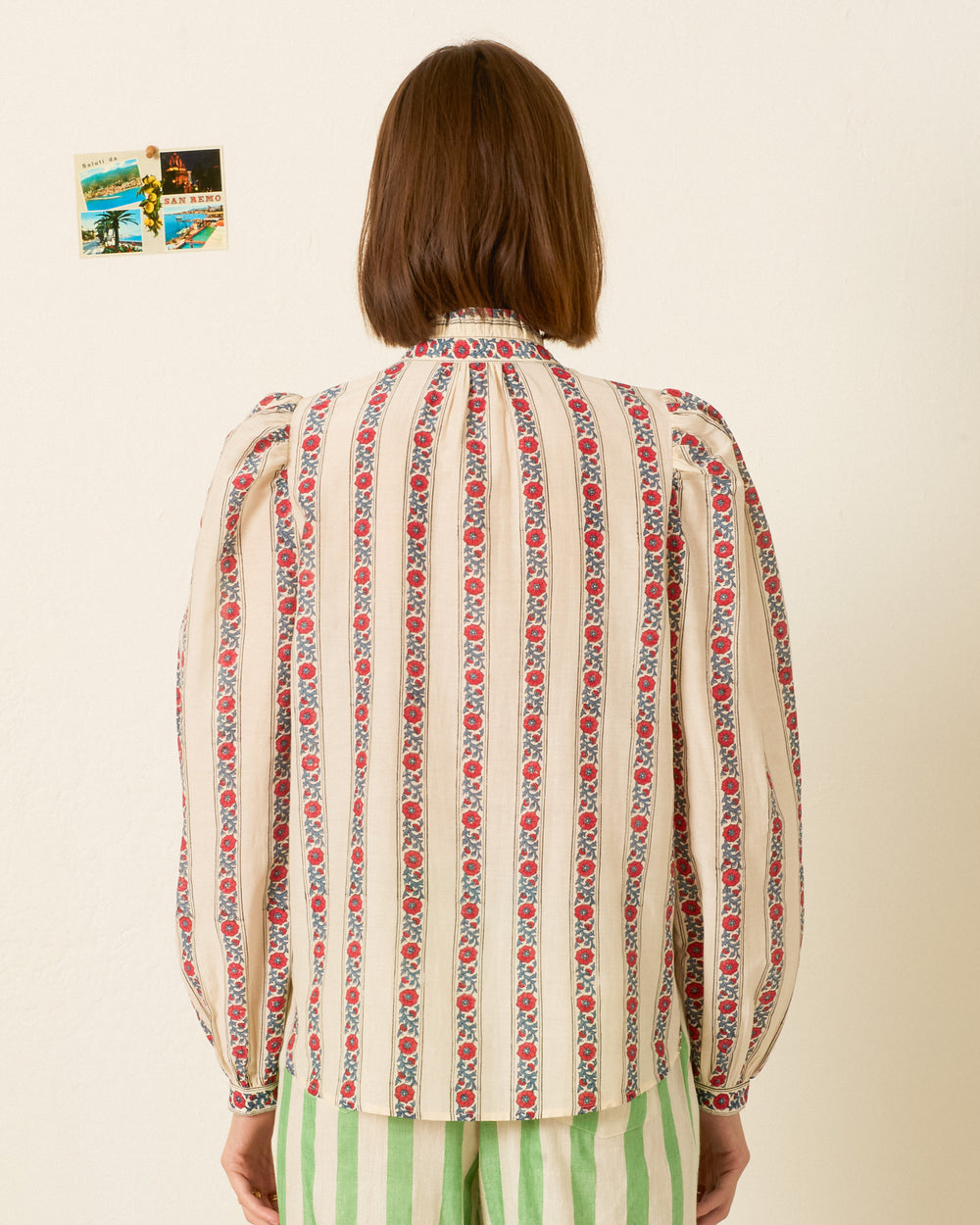 A person with straight brown hair faces away, wearing the Annabel Roseband Stripe Shirt—a hand-block-printed, long-sleeve cotton-silk blend top featuring red and blue vertical floral stripes and a subtle ruffled collar—paired with green striped pants. Two small postcards are visible on the wall in the background.