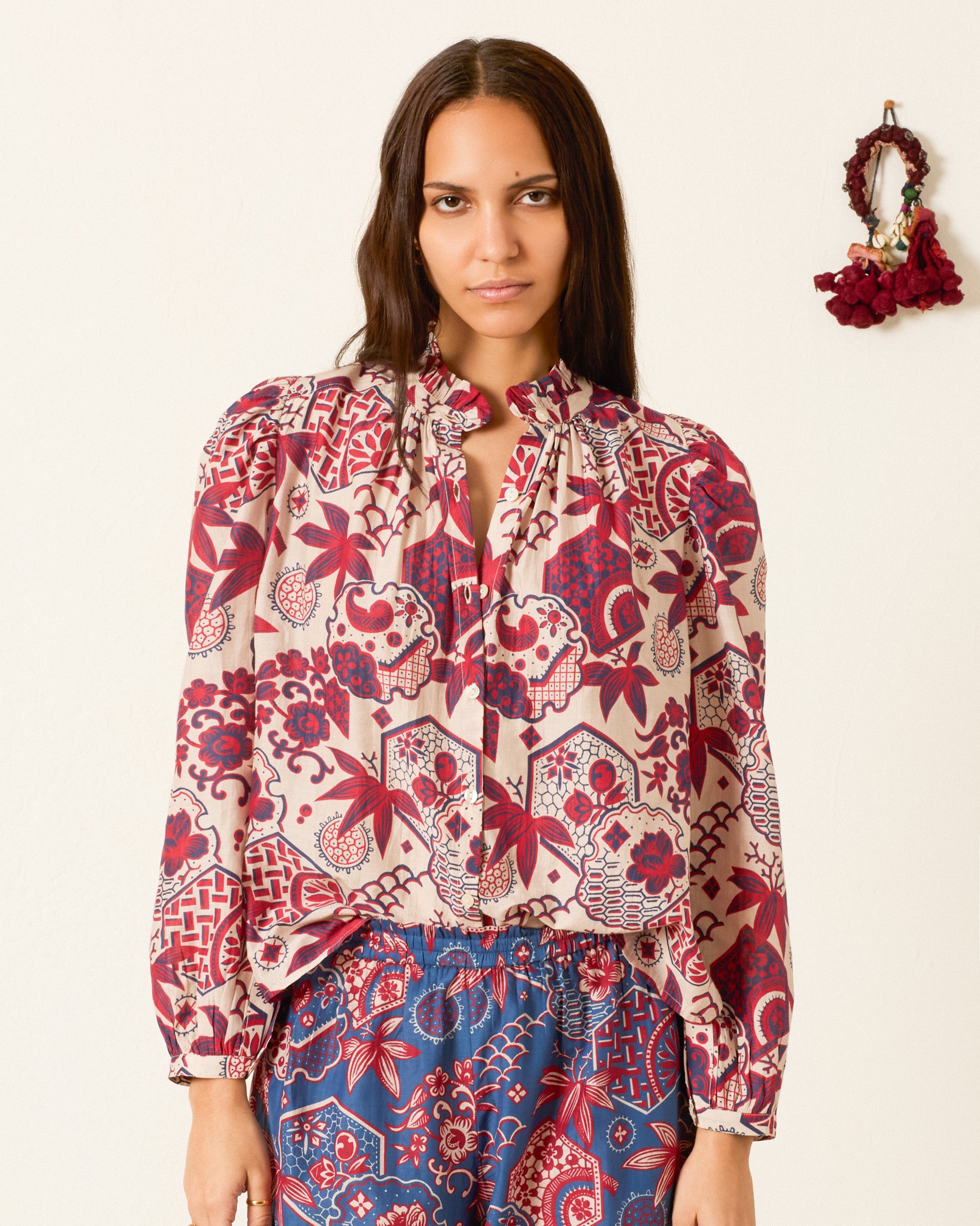 A woman wears the Annabel Crimson Hive Shirt, a vintage-inspired piece featuring red and blue designs and crafted from a soft cotton-silk blend, as she stands against a plain wall adorned with a small dried flower wreath.
