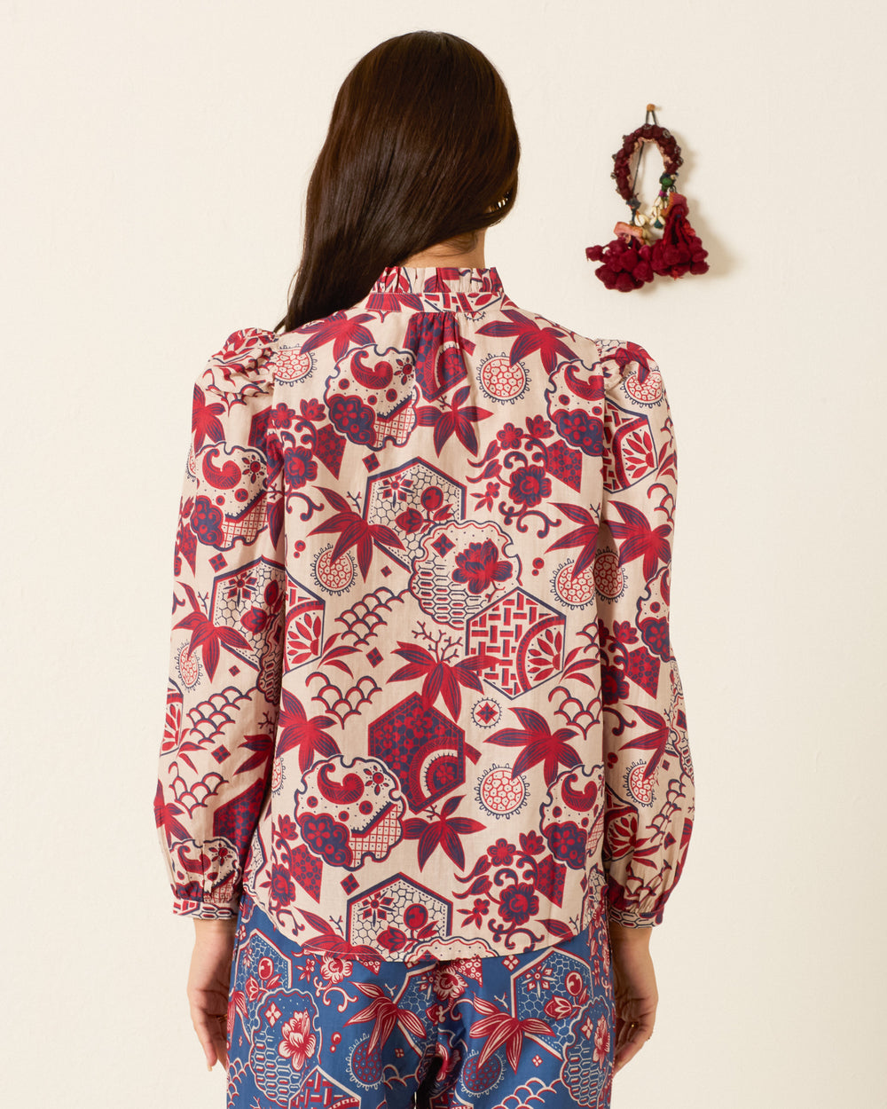A woman with long brown hair, standing with her back to the camera, wears the Annabel Crimson Hive Shirt— a red and beige patterned blouse featuring a ruffled collar—paired with blue patterned pants. A small decorative wreath hangs on the wall.