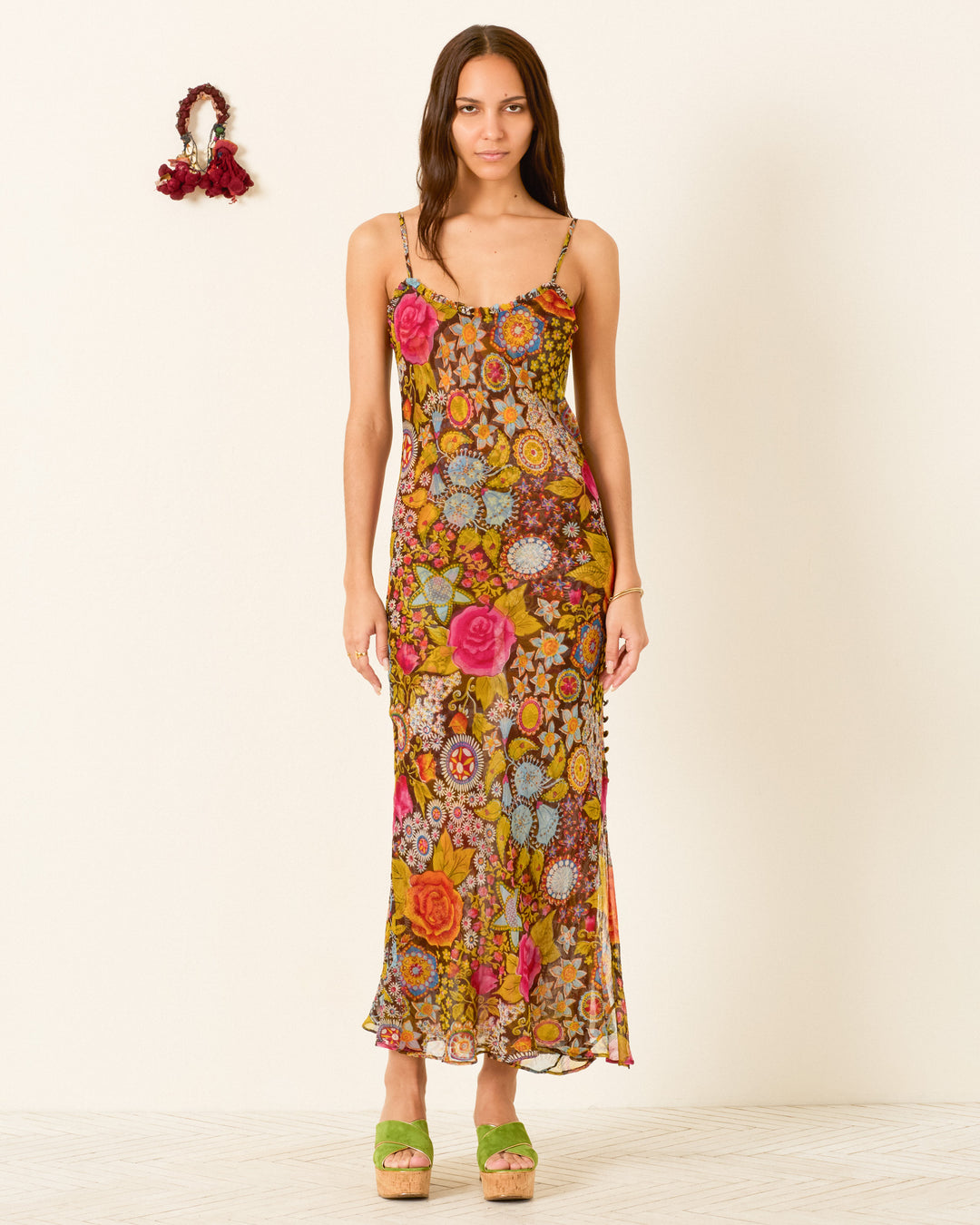 A woman stands against a plain background wearing the Athens Bloom Mirage Dress, a floral-patterned, sleeveless maxi with a ruffle-trimmed neckline, paired with green platform sandals.