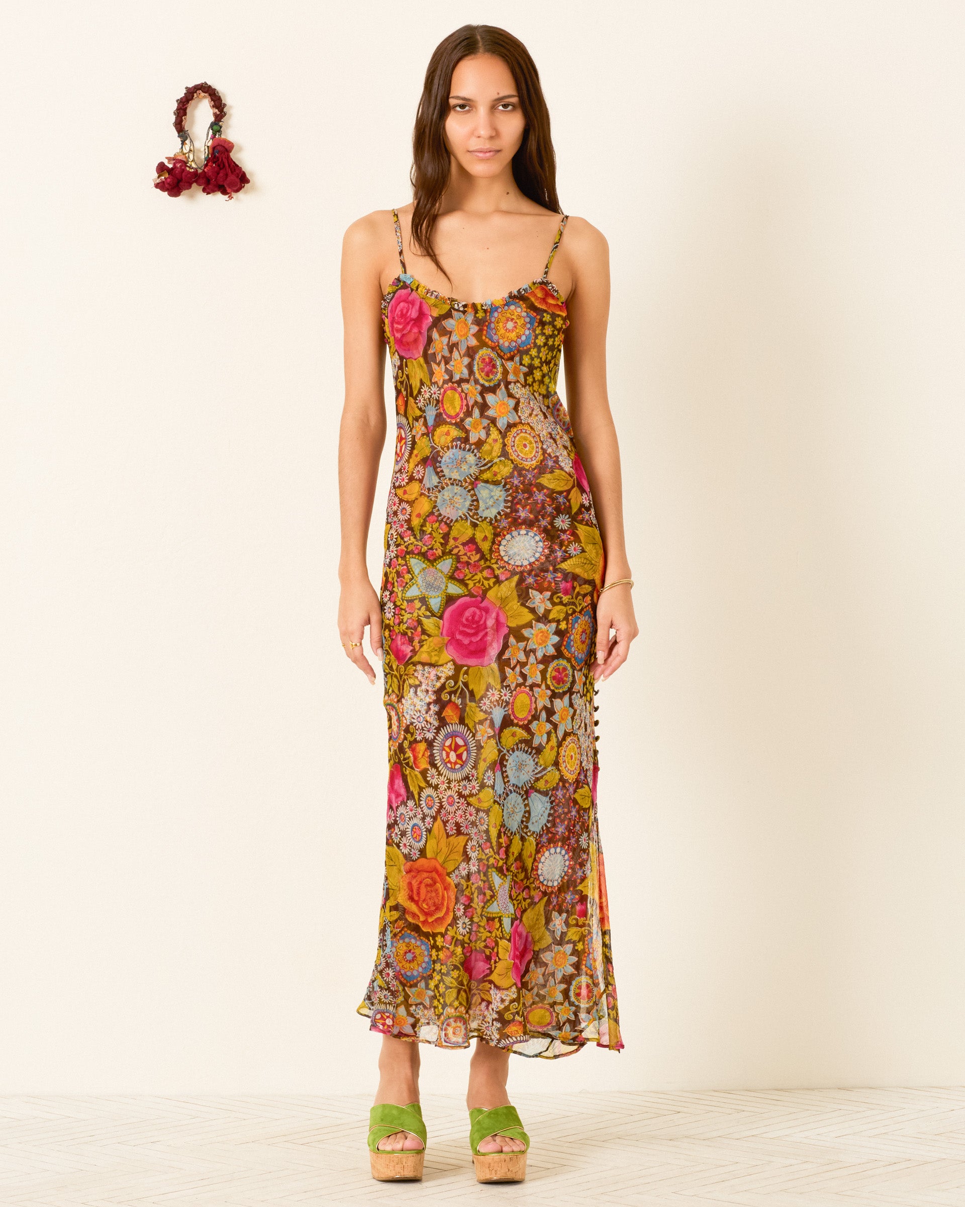 A woman stands against a plain background wearing the Athens Bloom Mirage Dress, a floral-patterned, sleeveless maxi with a ruffle-trimmed neckline, paired with green platform sandals.