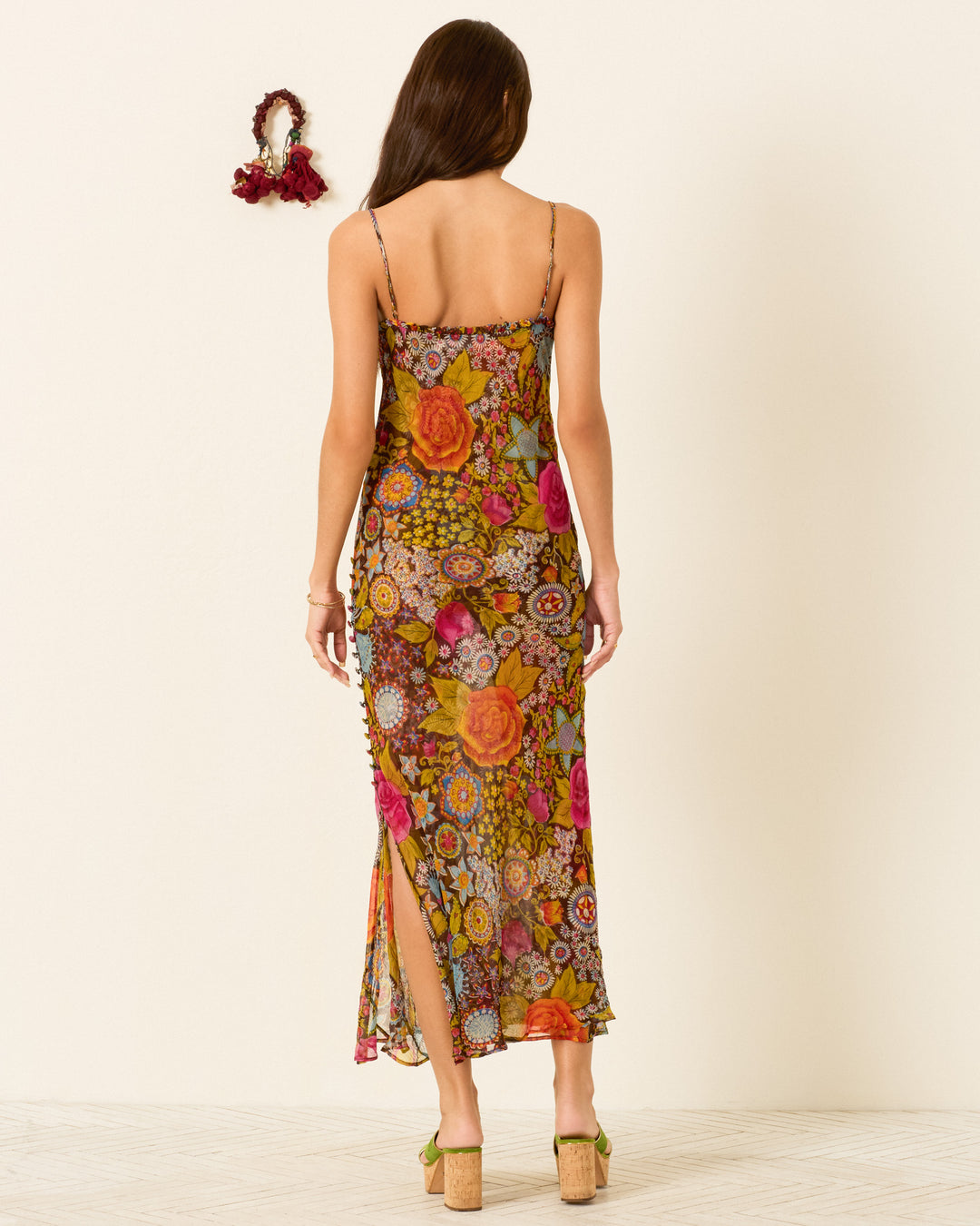 A woman faces away indoors, wearing the Athens Bloom Mirage Dress—a colorful strappy dress featuring a floral motif, ruffle-trimmed neckline, and side slit. A floral hair accessory hangs on the wall nearby.