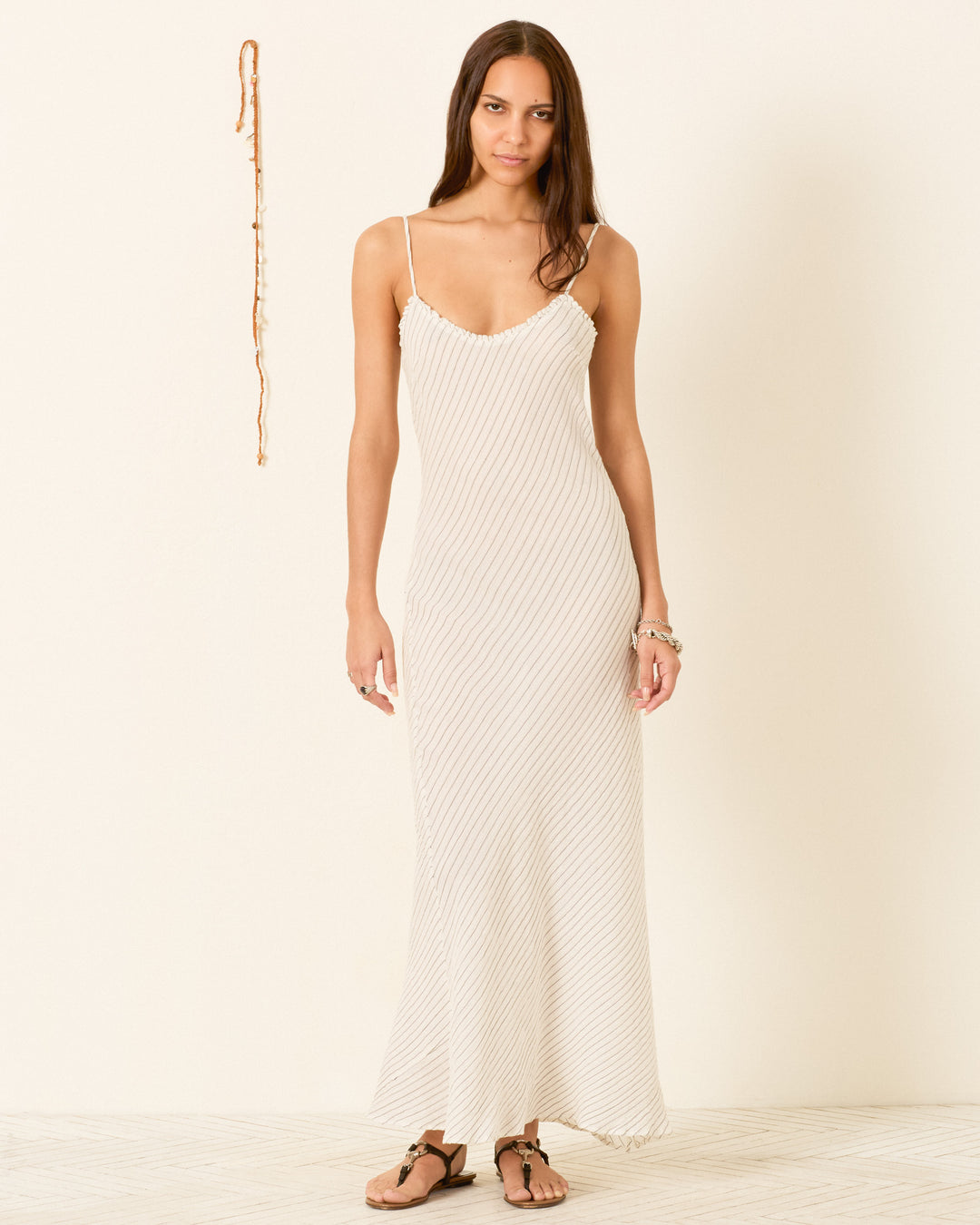 A woman stands indoors against a plain wall, wearing the Athens Sea Salt Dress—a long, fitted slip dress with spaghetti straps and subtle yarn-dyed stripes—styled with bracelets and sandals.