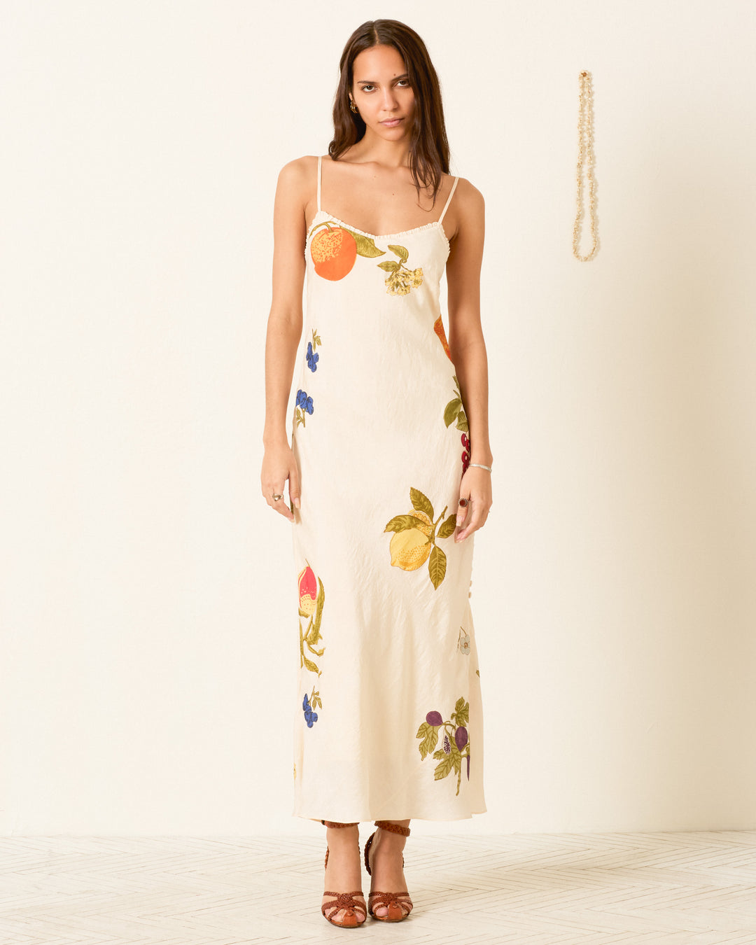 A woman wearing the Athens Nine Fruit Silk Dress—a cream-colored, bias-cut slip dress featuring fruit and floral prints—stands against a plain wall with a pearl necklace hanging beside her.