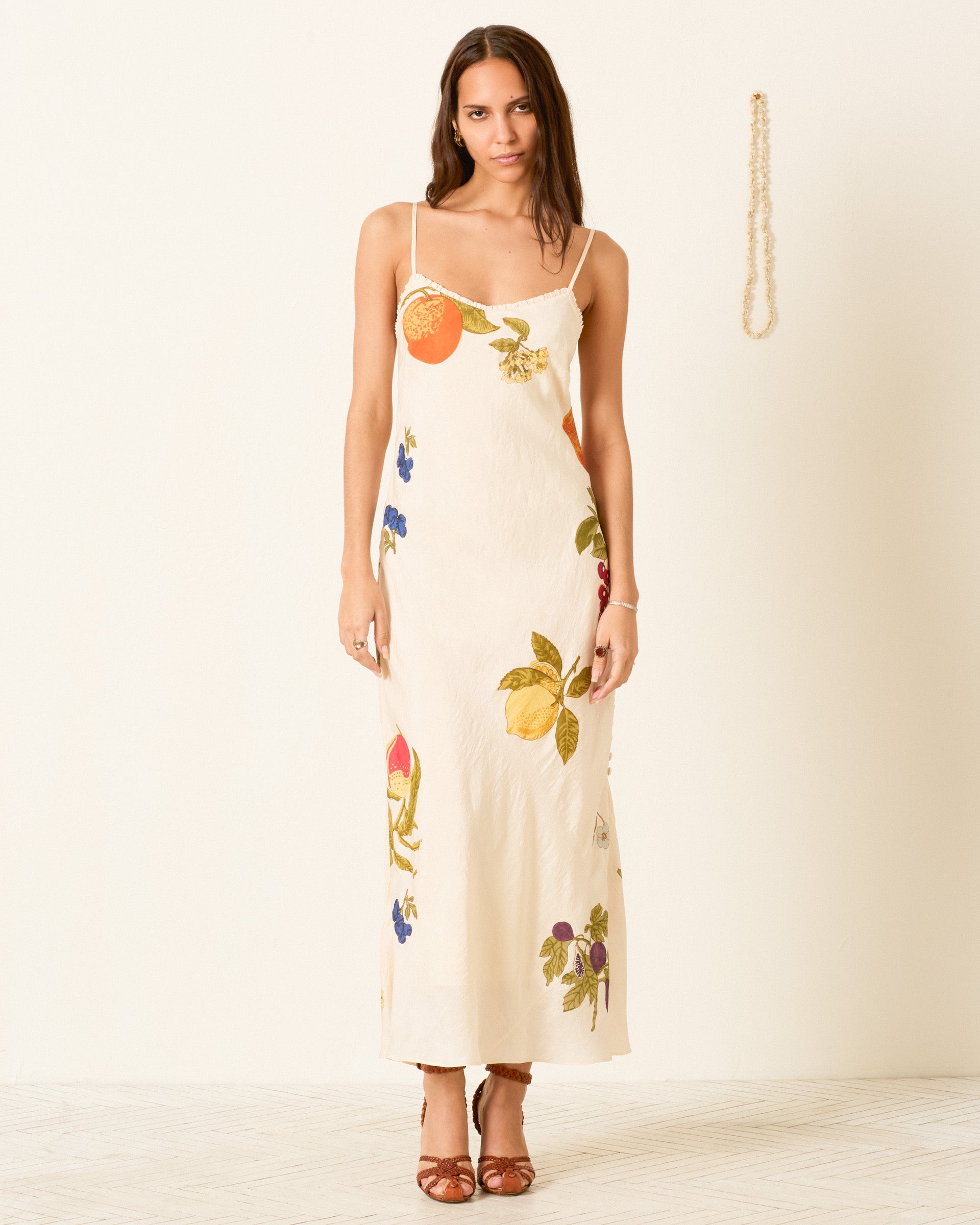 A woman wearing the Athens Nine Fruit Silk Dress—a cream-colored, bias-cut slip dress featuring fruit and floral prints—stands against a plain wall with a pearl necklace hanging beside her.