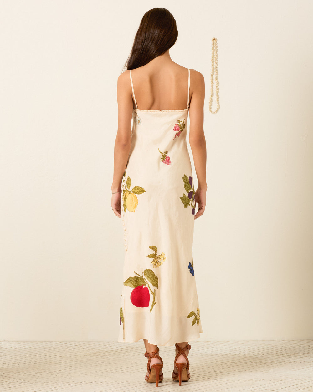 A woman stands with her back to the camera wearing the Athens Nine Fruit Silk Dress—a hand-block-printed, cream-colored piece adorned with colorful fruit and floral motifs, featuring spaghetti straps. She pairs the dress with brown sandal heels, while a pearl necklace hangs on the wall to her right.