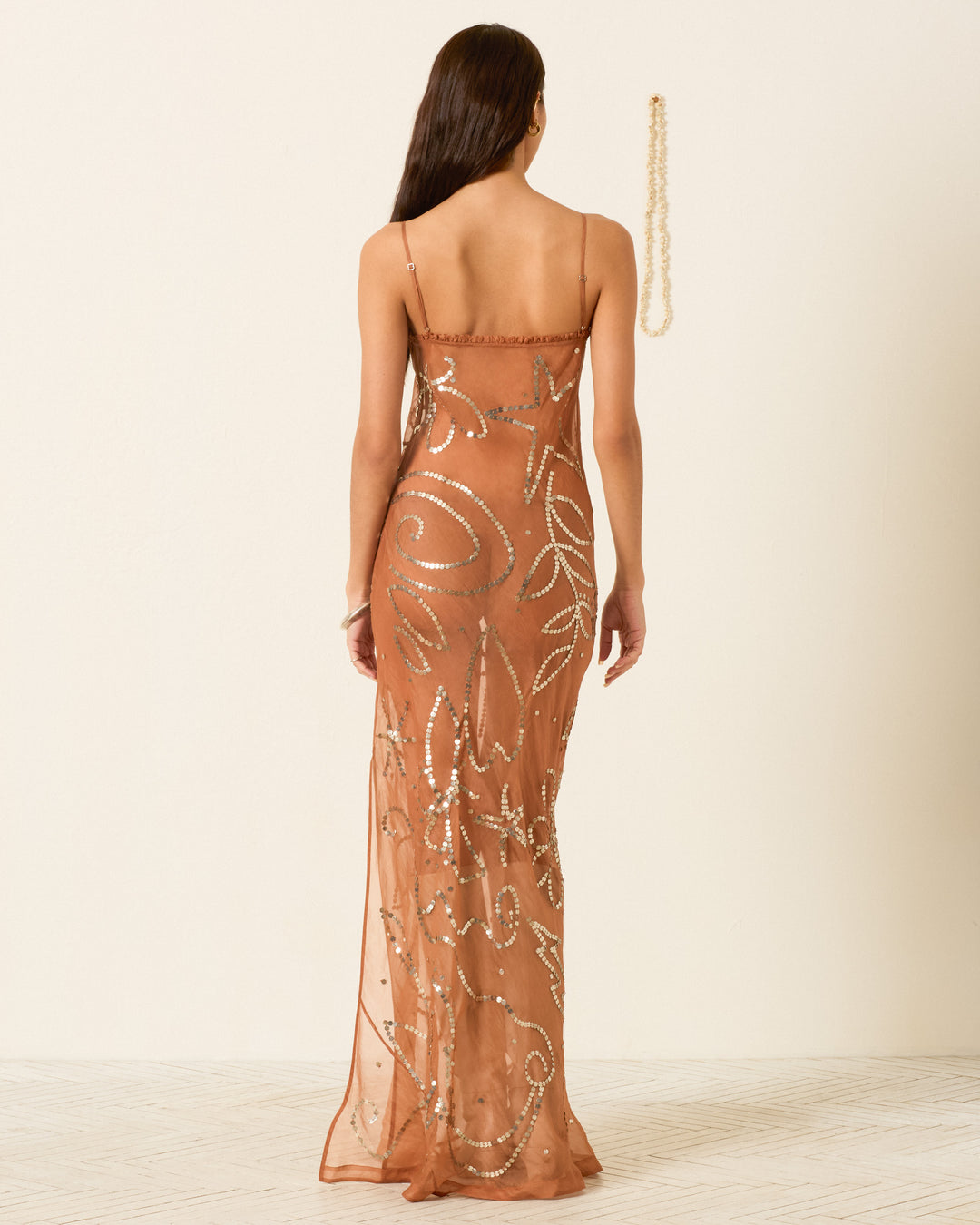 A woman with long brown hair faces away from the camera, wearing the Athens Fawn Silk Coin Dress—a sheer, hand-embellished, sequined floor-length gown with thin straps and a ruffle-trimmed neckline—against a plain light background.