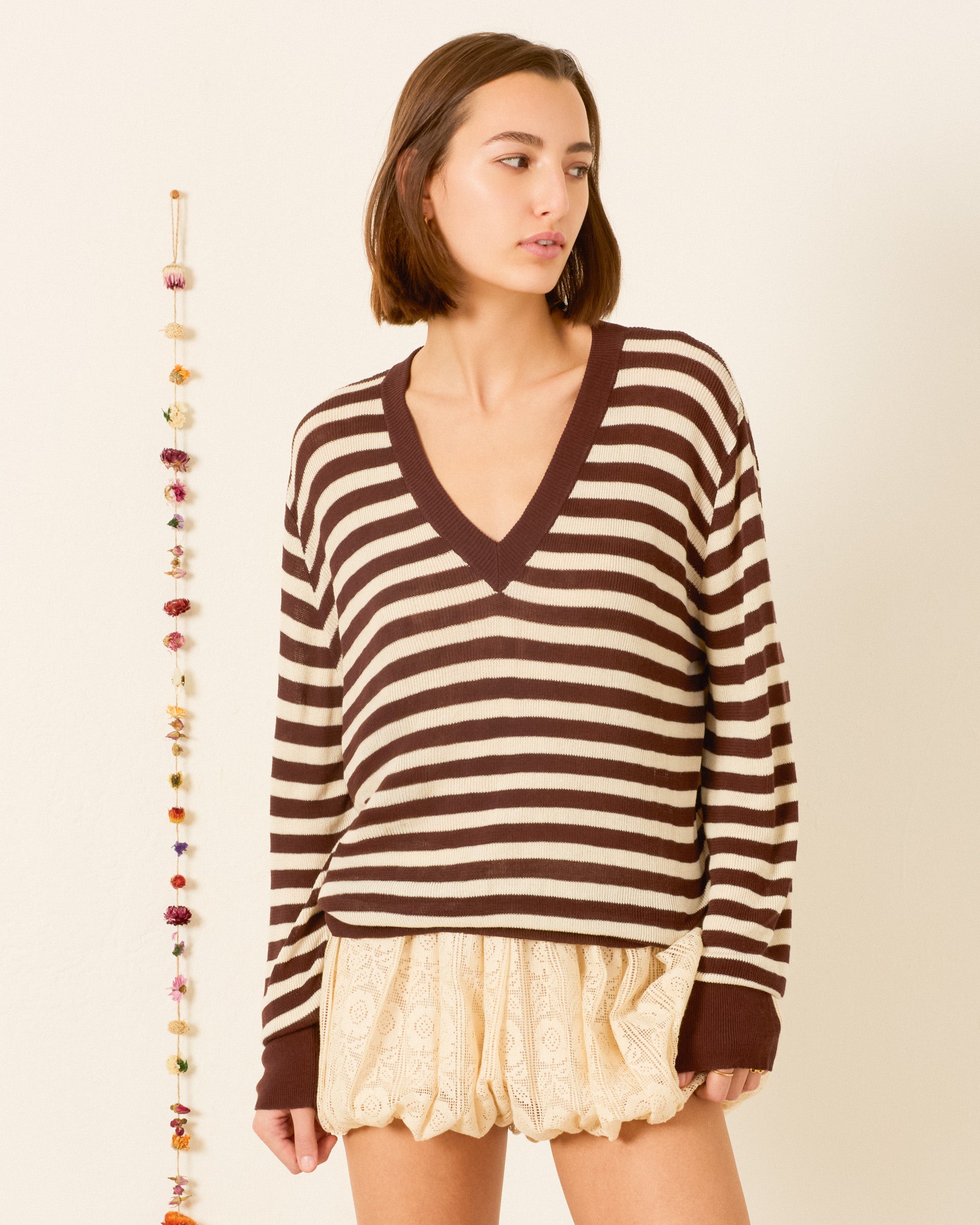 A woman wearing the Axel Clove Stripe Sweater, featuring brown and beige stripes, stands against a plain background with a hanging garland beside her.