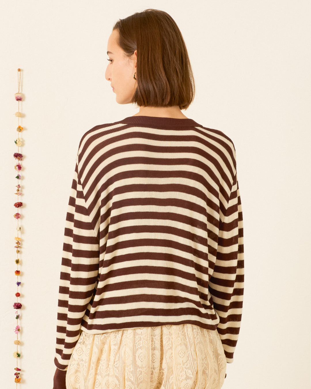 A person with short brown hair is shown from the back, wearing the Axel Clove Stripe Sweater in soft bamboo viscose and light-colored textured pants, standing near a string of hanging decorations.