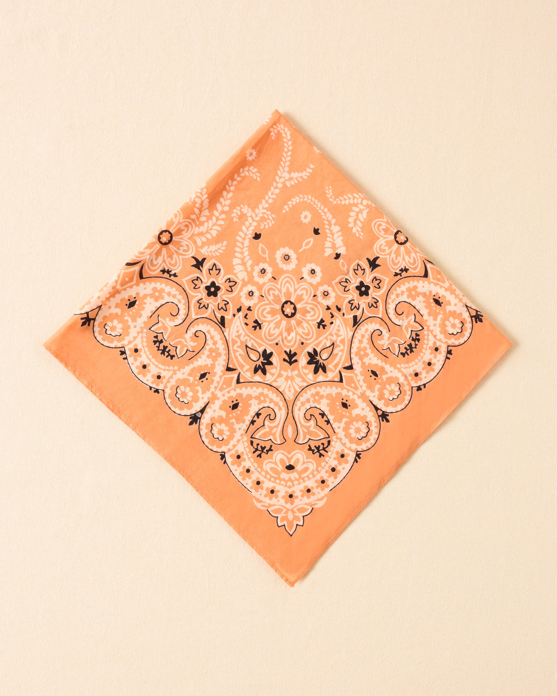 The Bodhi August Bandana, in a peach cotton-silk blend with a white and black botanical motif, is displayed flat on a light beige surface.