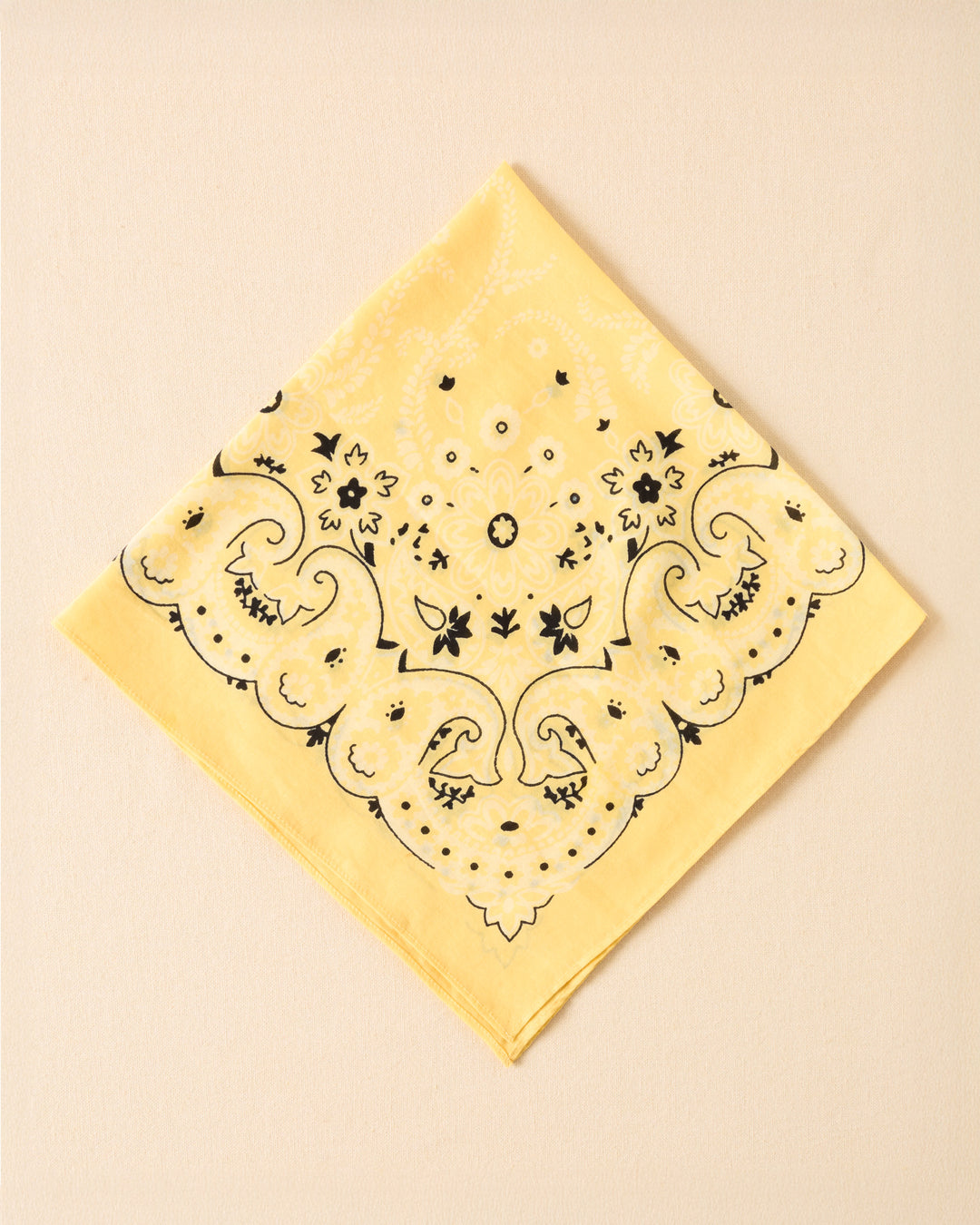The Bodhi Sunlight Bandana, featuring a yellow base with a black and white paisley pattern, is neatly folded into a square and set against a plain beige background.